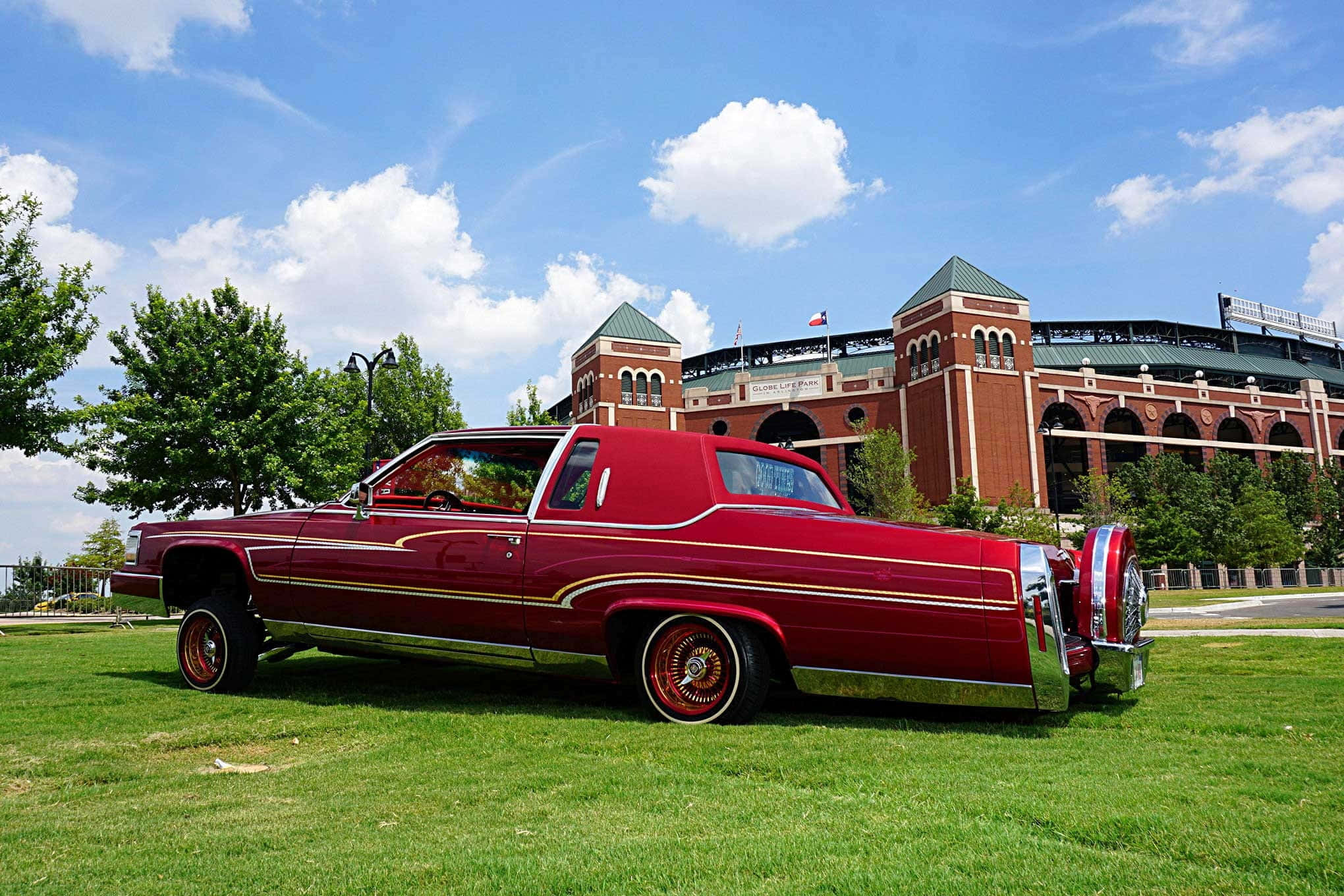 Cadillac Fleetwood 桌布 壁紙 — 一輛閃亮的紅色經典汽車帶有金色飾邊，停在綠色草地上，前面是一個大型磚砌體育場，上方是晴朗的藍天和蓬鬆的白雲。