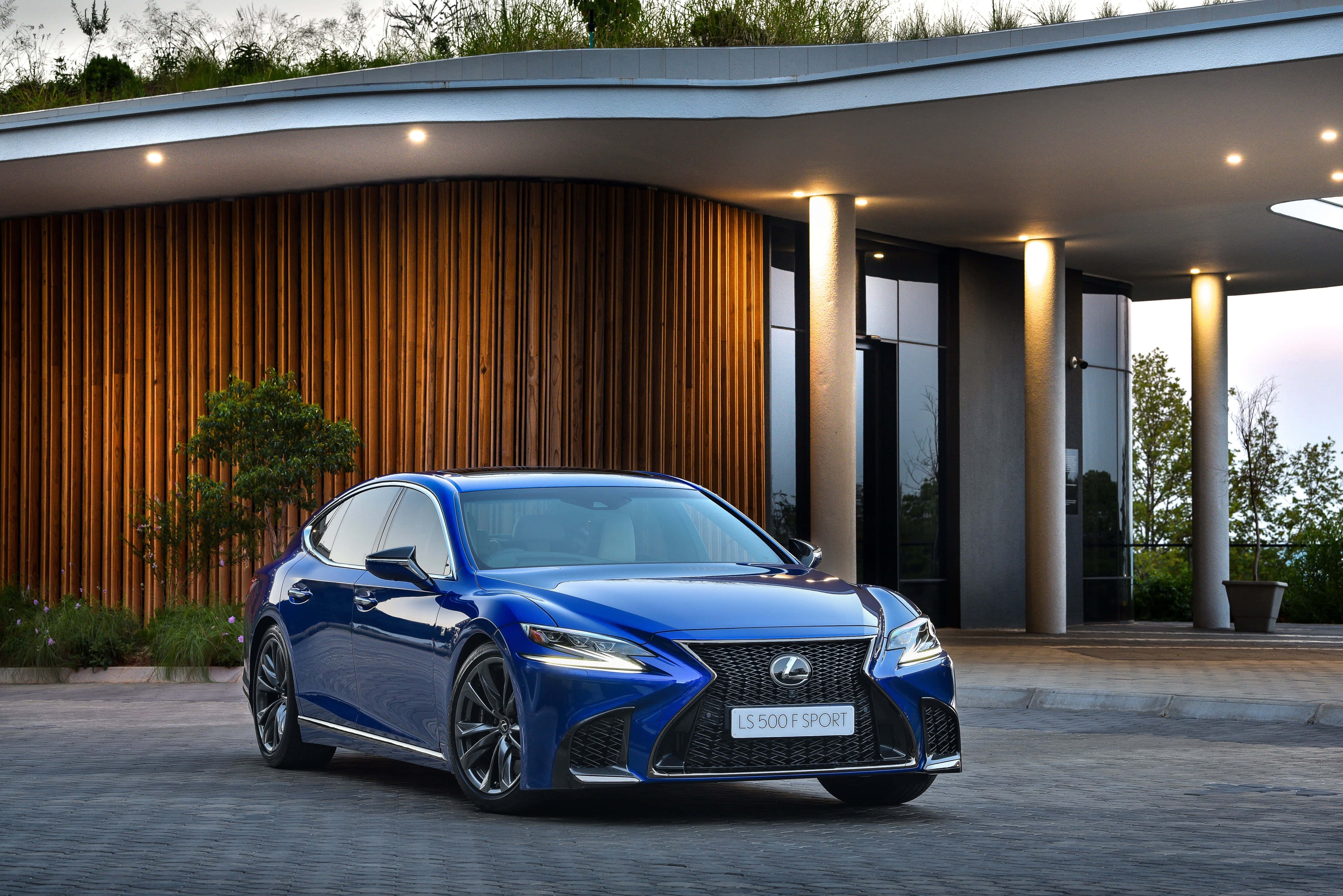 Lexus Ls 500 桌布 壁紙 — 一輛閃亮的藍色Lexus LS 500 F Sport優雅地停在現代木質外牆房屋前，配有環境照明。