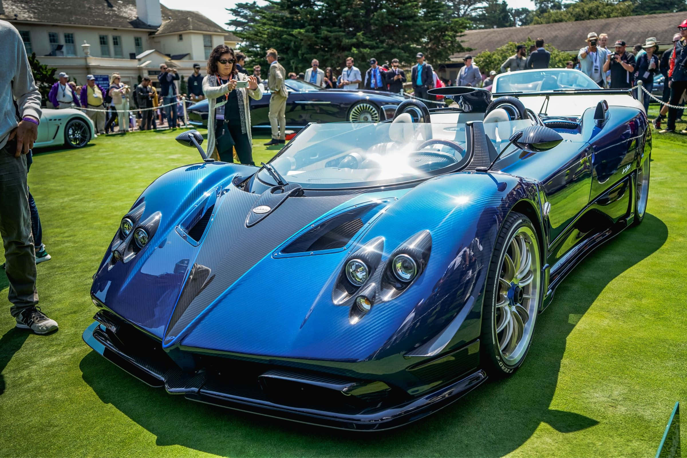 Pagani Zonda Hp Barchetta 桌布 壁紙 — 一輛閃亮的藍色帕加尼 Huayra 跑車在戶外車展的修剪整齊的綠色草坪上，在明亮的陽光下閃閃發光。