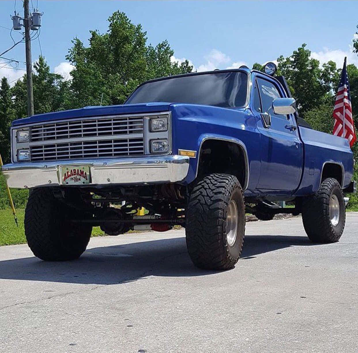 Square Body Truck 桌布 壁紙 — 一輛升高了的藍色經典雪佛蘭皮卡車，帶有阿拉巴馬州車牌和美國國旗，自豪地站在晴朗藍天下的柏油路上。