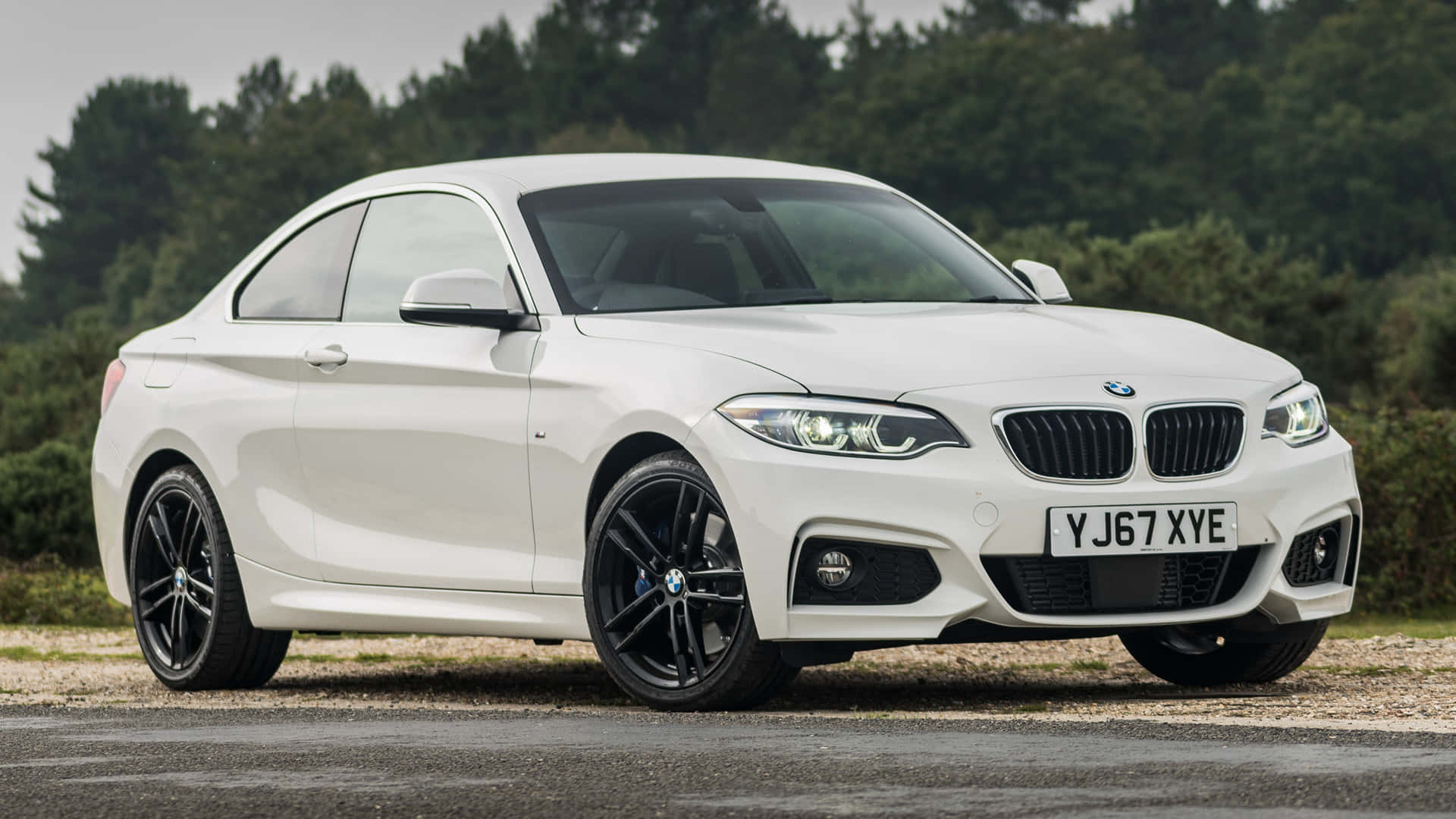 Bmw 2 Series 桌布 壁紙 — 一輛時尚的白色BMW 2系轎跑車，配黑色輪圈，停泊在碎石路上，旁邊是樹林，天空多雲。
