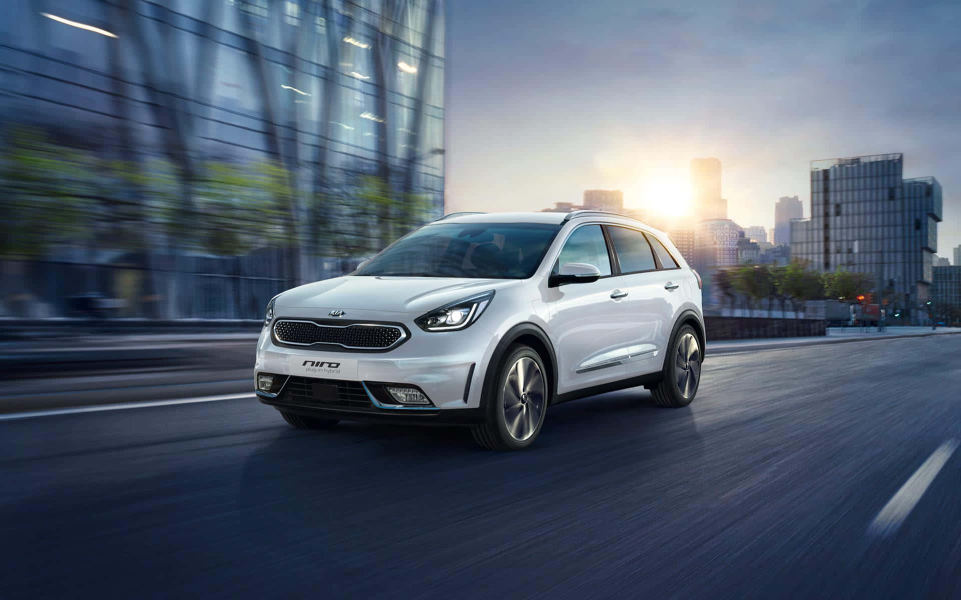 Kia Niro 桌布 壁紙 — 一輛時尚的白色Kia Niro SUV在日落時分沿著城市高速公路疾馳，背景有動態模糊和發光的摩天大樓。