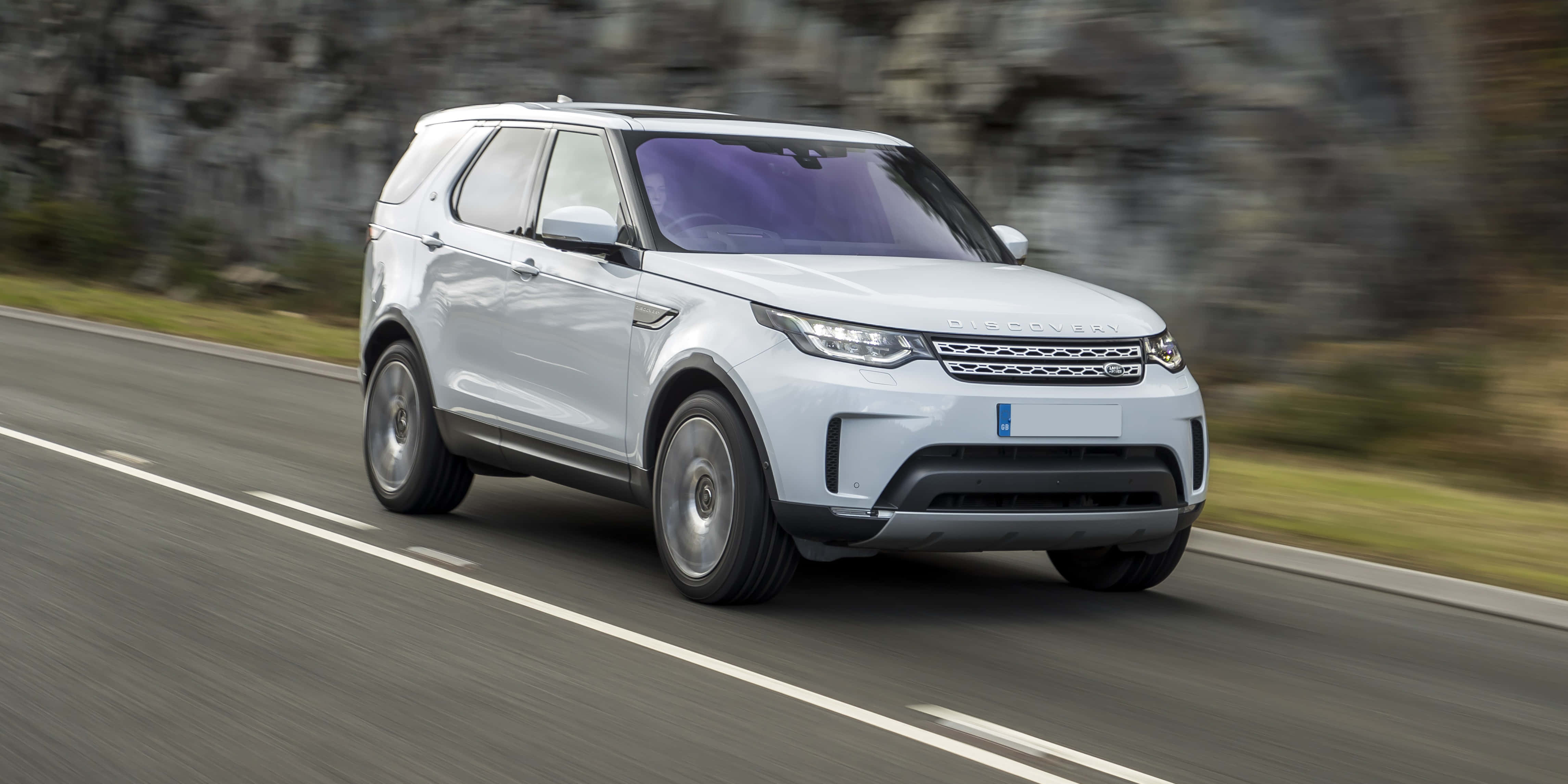 Land Rover Discovery 桌布 壁紙 — 一輛時尚的白色SUV在蜿蜒的道路上行駛，背景是模糊的岩石懸崖和綠植。