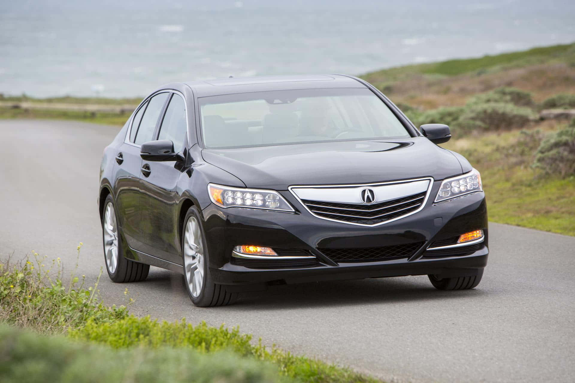 Acura Rlx 桌布 壁紙 — 一輛時尚的黑色Acura轎車沿著海岸道路行駛，背景是海洋和綠色灌木。