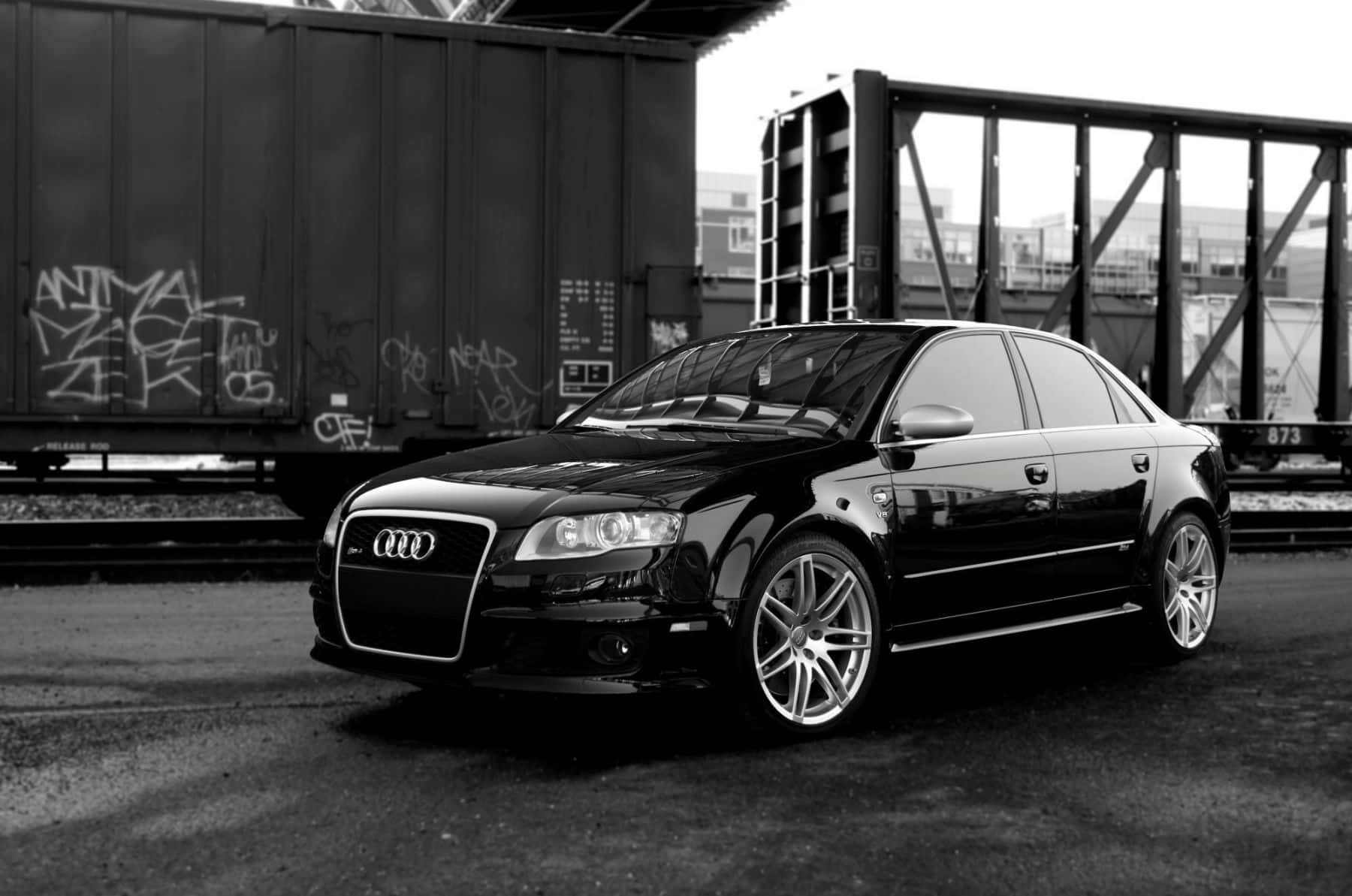 Audi Rs4 桌布 壁紙 — 一輛時尚的黑色奧迪轎車配備定制輪圈，停在工業橋下塗滿塗鴉的火車車廂旁。