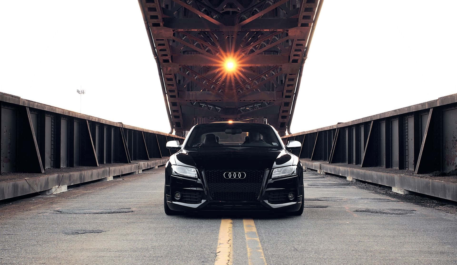 Audi S6 桌布 壁紙 — 一輛時尚的黑色奧迪跑車在鋼橋下向前，明亮的太陽在上方的拱門中閃耀。
