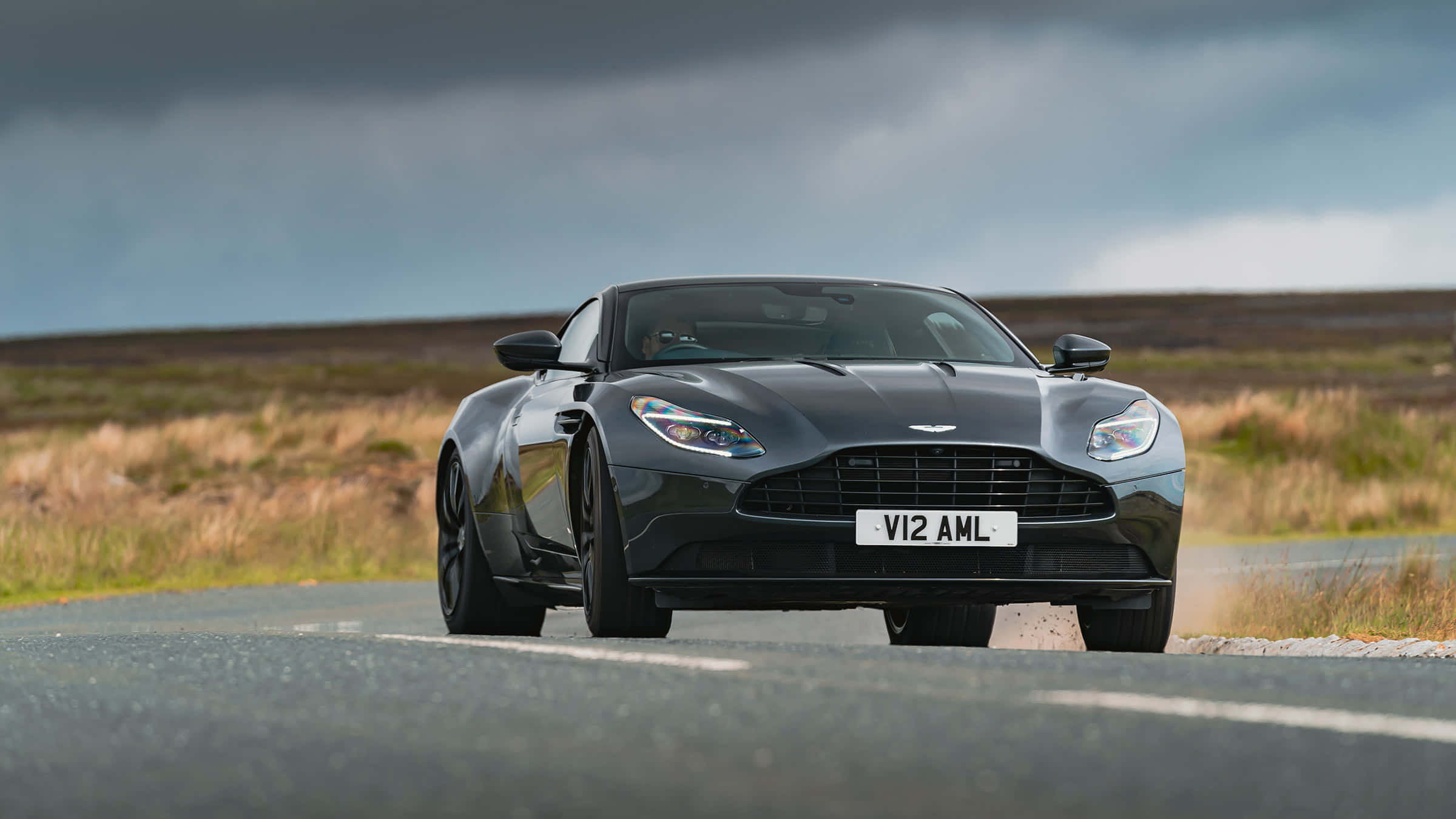 Aston Martin Db11 桌布 壁紙 — 一輛時尚的黑色Aston Martin V12 AMR在戲劇性的多雲天空下沿鄉村道路飛馳。