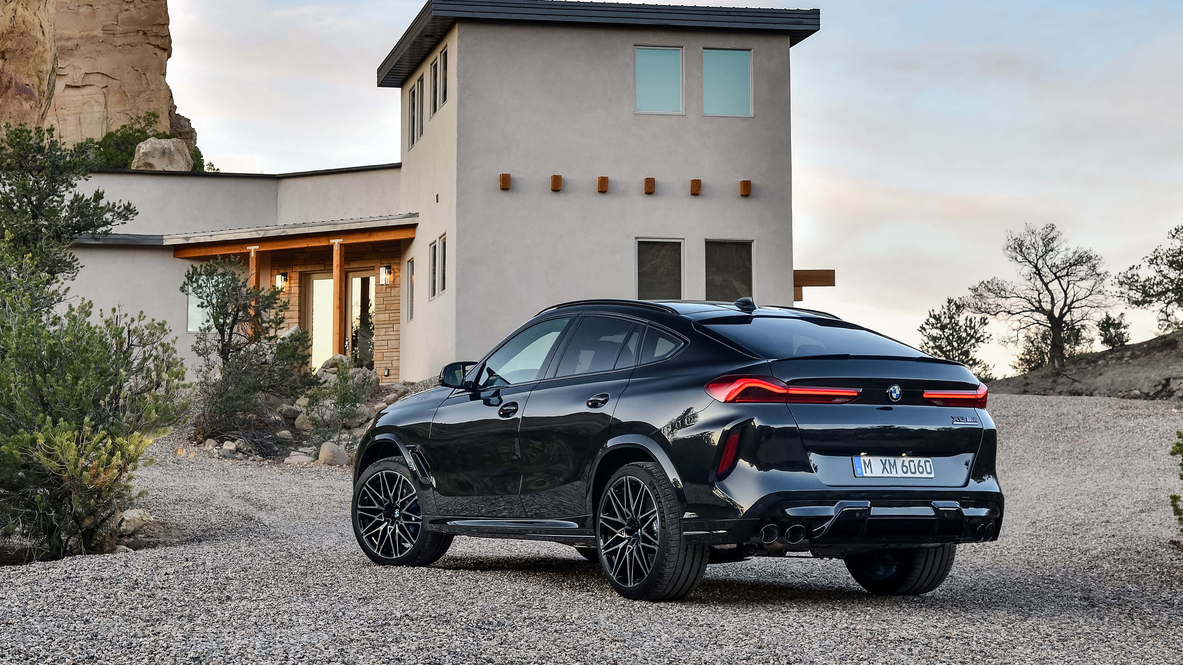 Bmw X6 桌布 壁紙 — 一輛時尚的黑色BMW X6 M停在碎石路上，前面是現代沙漠房屋，周圍有岩石地貌和稀疏樹木。