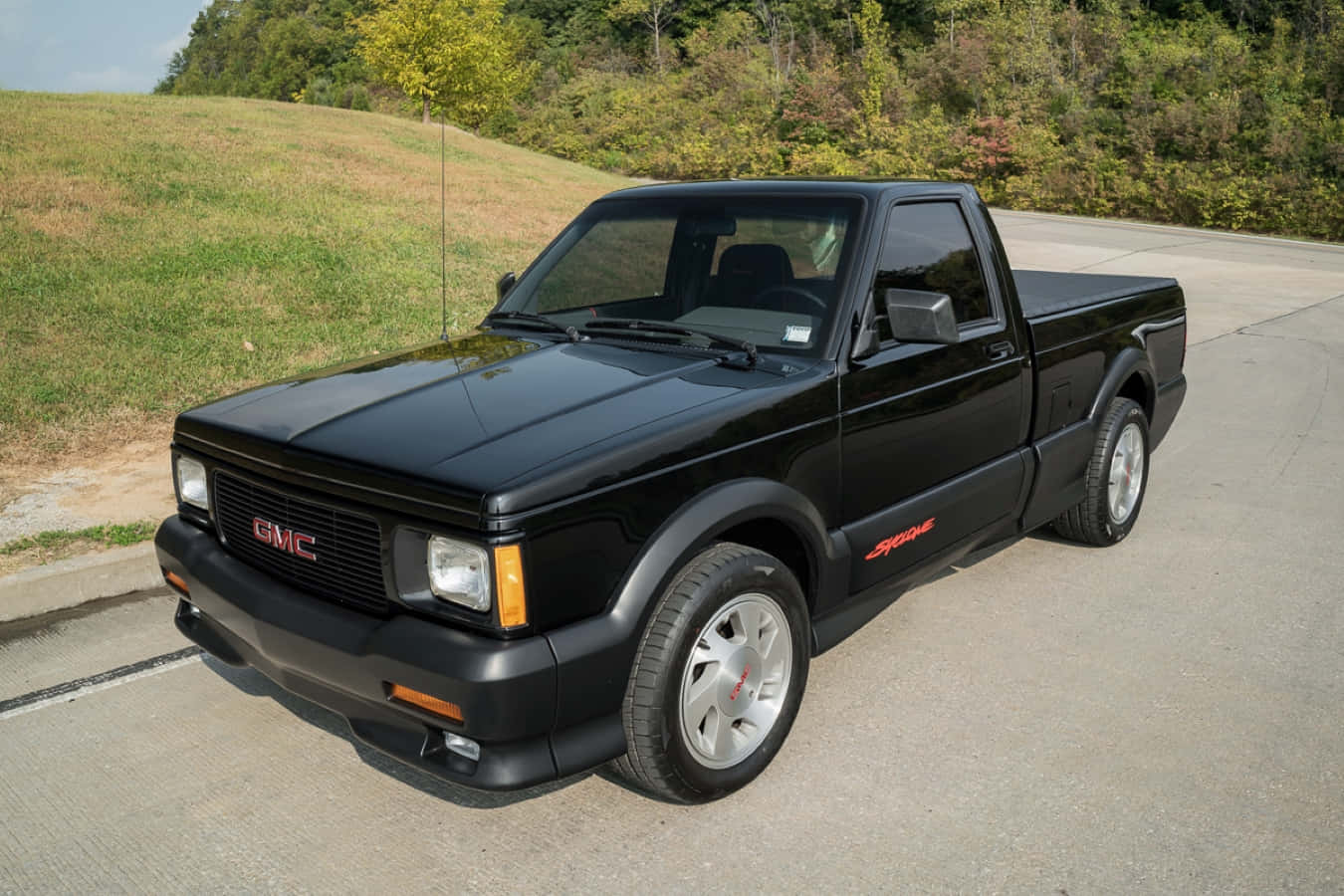 Gmc Syclone 桌布 壁紙 — 一輛時尚的黑色GMC Syclone pickup貨車停泊在鋪砌道路上，旁邊是長滿秋樹的草坡。