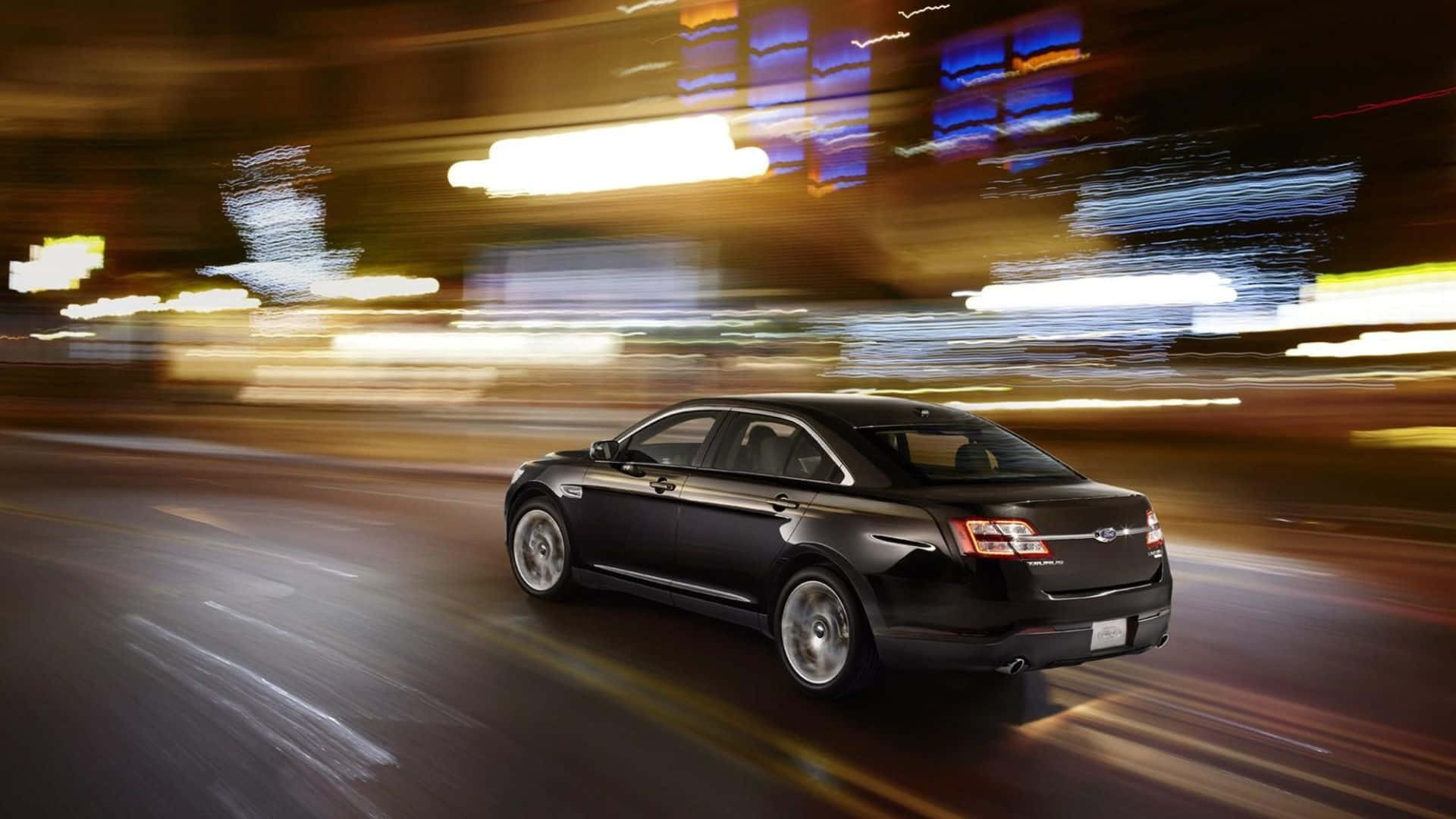 Ford Taurus 桌布 壁紙 — 一輛時尚的黑色轎車在夜晚飛馳於霓虹燈閃爍的城市中，燈光拖曳模糊了都市背景。