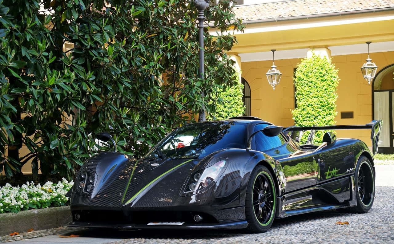 Pagani Zonda Tricolore 桌布 壁紙 — 一輛時尚的黑色Pagani Zonda跑車，帶有綠色點綴，停在鵝卵石車道上，旁邊是茂密的綠植和一座黃色別墅。