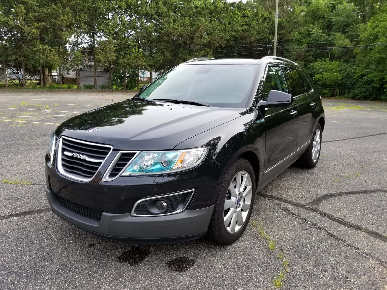 Saab 9-4x 桌布 壁紙 — 一輛時尚的黑色薩博SUV停在一個佈滿裂縫的瀝青停車場，周圍環繞著茂密的綠樹。
