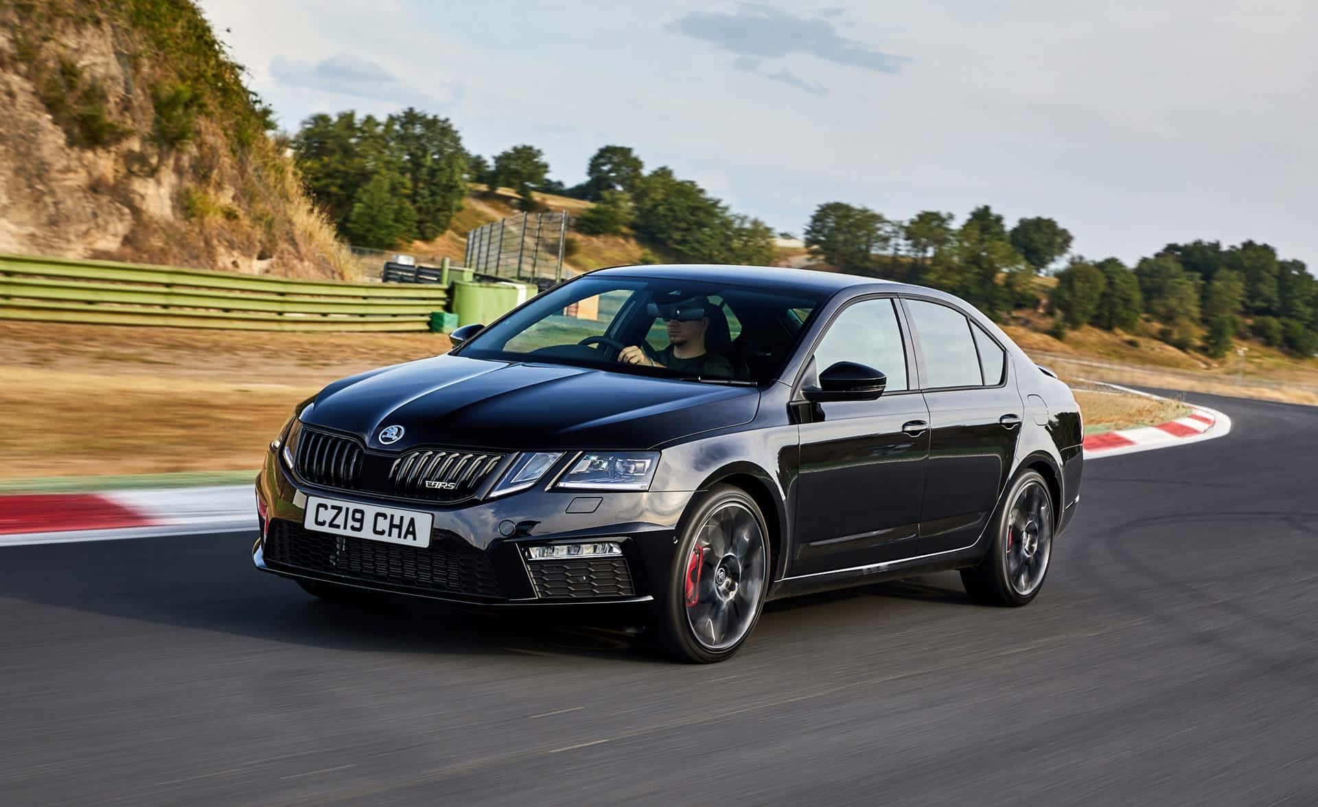 Skoda Octavia 桌布 壁紙 — 一輛時尚的黑色斯柯達明銳RS在部分多雲的天空下，繞著帶有紅色和白色路緣的賽道彎道飛馳。