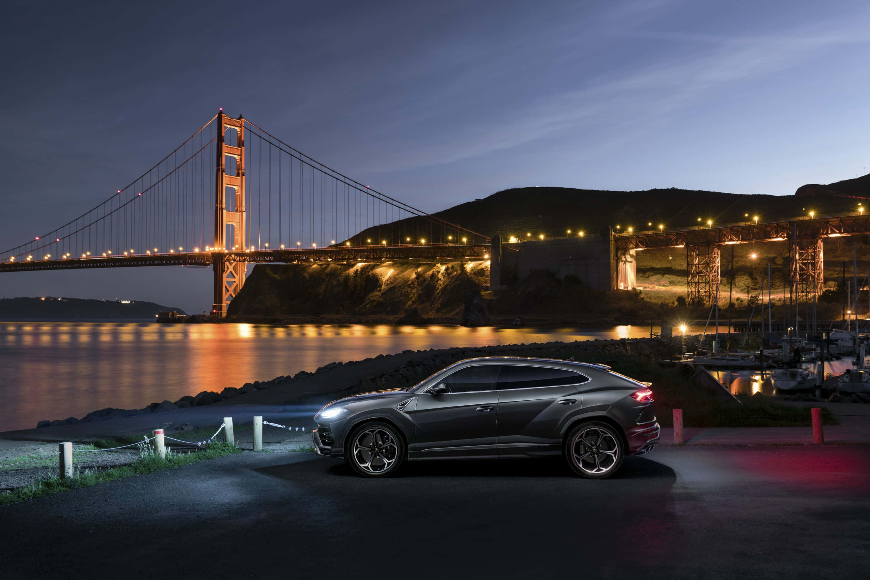 Lamborghini Urus 桌布 壁紙 — 一輛時尚的灰色SUV在黃昏時停泊，背後的金門大橋亮著燈光，在水面上閃耀。