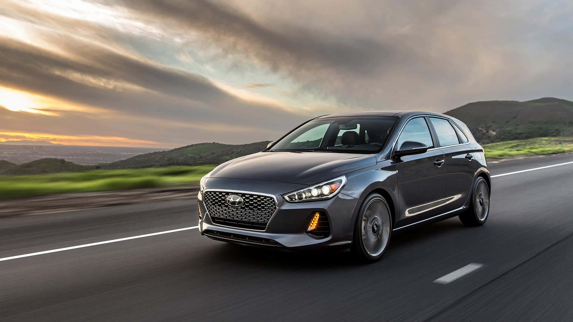 Hyundai Elantra 桌布 壁紙 — 一輛時尚的灰色現代 i30 掀背車在日落時分沿著蜿蜒的道路疾馳，背景是起伏的山丘和戲劇性的雲彩。
