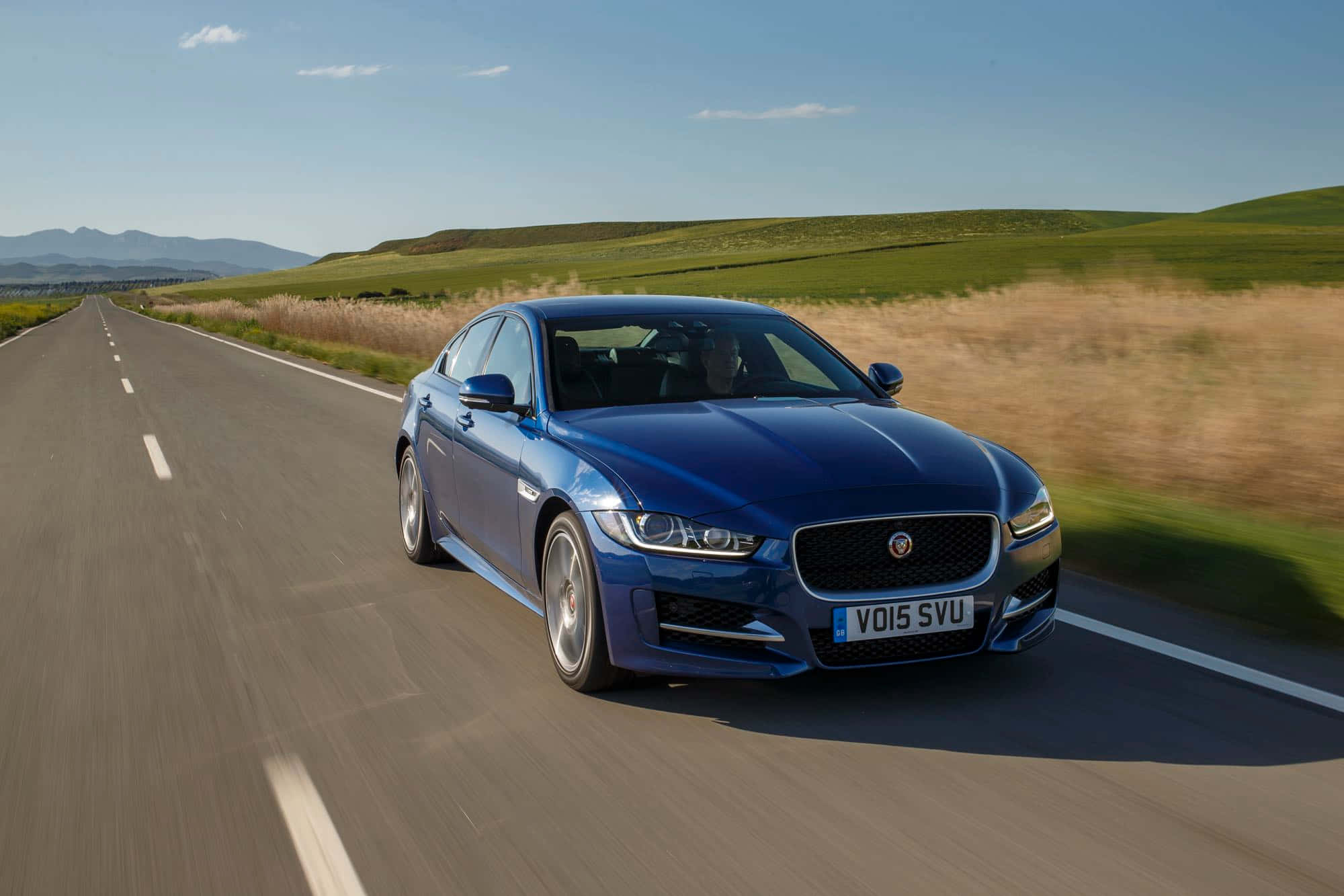 Jaguar Xe 桌布 壁紙 — 一輛時尚的藍色積架XF房車在陽光明媚的鄉村高速公路上飛馳，沿途有連綿起伏的綠色山丘和遠處的山脈，天空清澈湛藍。