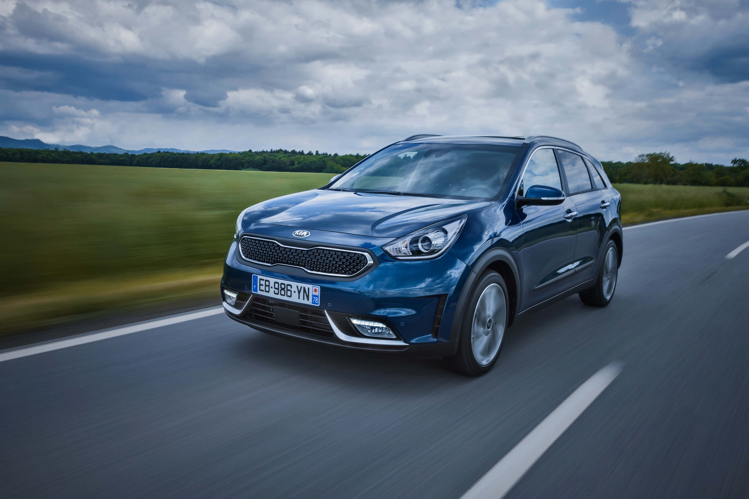 Kia Niro 桌布 壁紙 — 一輛時尚的藍色Kia SUV在鄉村道路上疾馳，背景是模糊的綠色田野和多雲的天空。