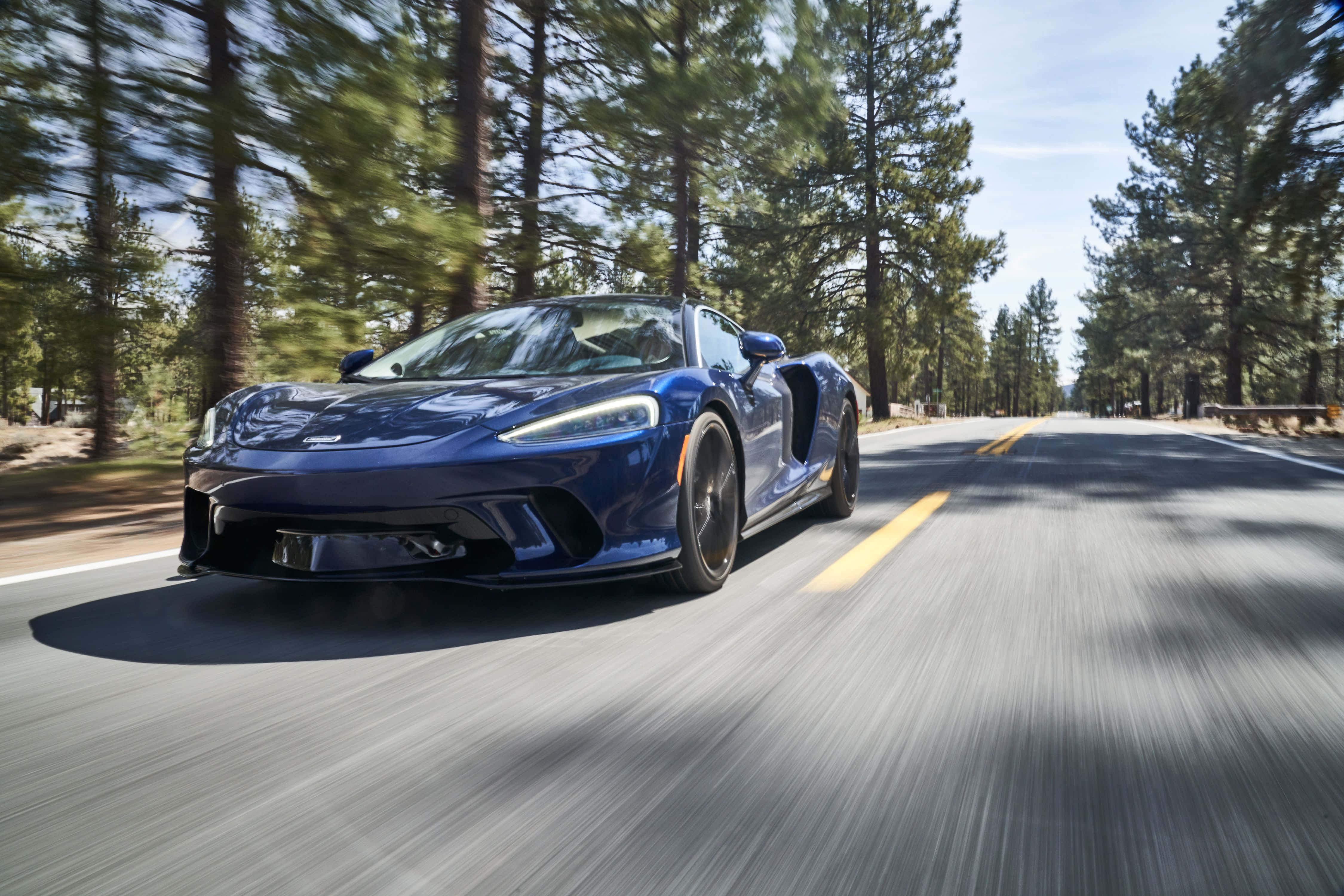 Mclaren Gt 桌布 壁紙 — 一輛時尚的藍色跑車在樹林環繞的道路上飛馳，動態模糊強調了速度。