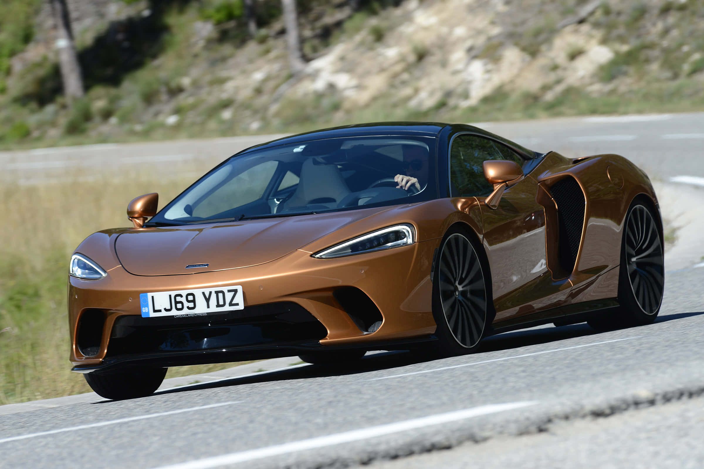 Mclaren Gt 桌布 壁紙 — 一輛時尚的青銅色McLaren跑車在明亮的日光下沿著蜿蜒的山路飛馳。