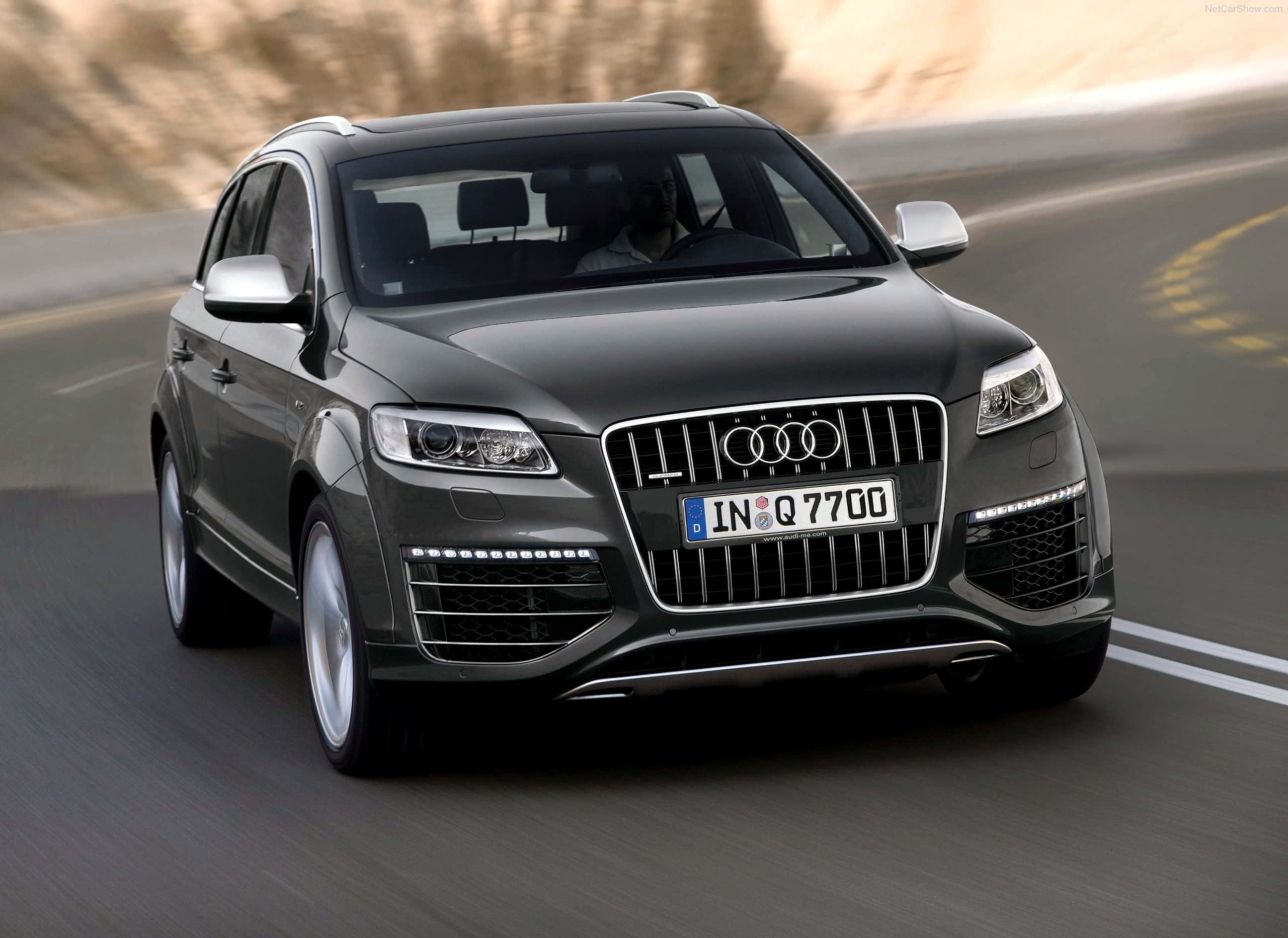 Audi Q7 桌布 壁紙 — 一輛時尚的深灰色奧迪Q7 SUV在蜿蜒的道路上行駛，背景有模糊的動態和陽光照耀的山丘。
