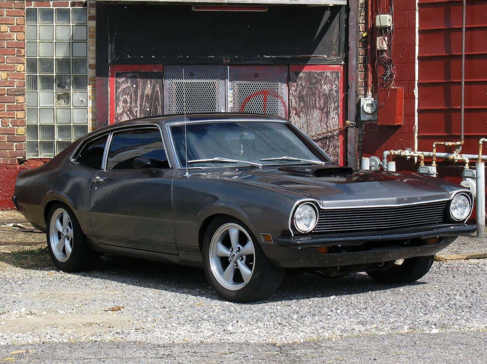 Ford Maverick 桌布 壁紙 — 一輛時尚的深灰色經典福特 Maverick 雙門轎跑車停在碎石路上，前面是一座帶有玻璃磚窗的風化紅磚建築。