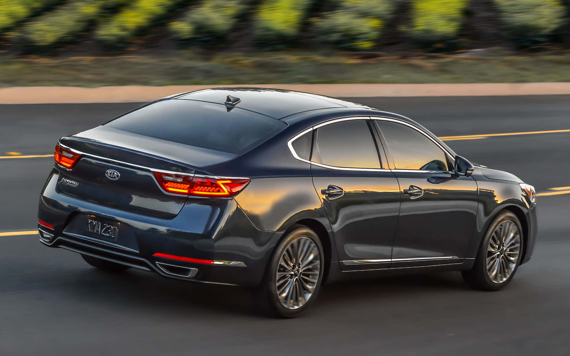 Kia Cadenza 桌布 壁紙 — 一輛時尚的深灰色Kia Optima房車在陽光照耀的道路上平穩行駛，背景是模糊的綠色植物。