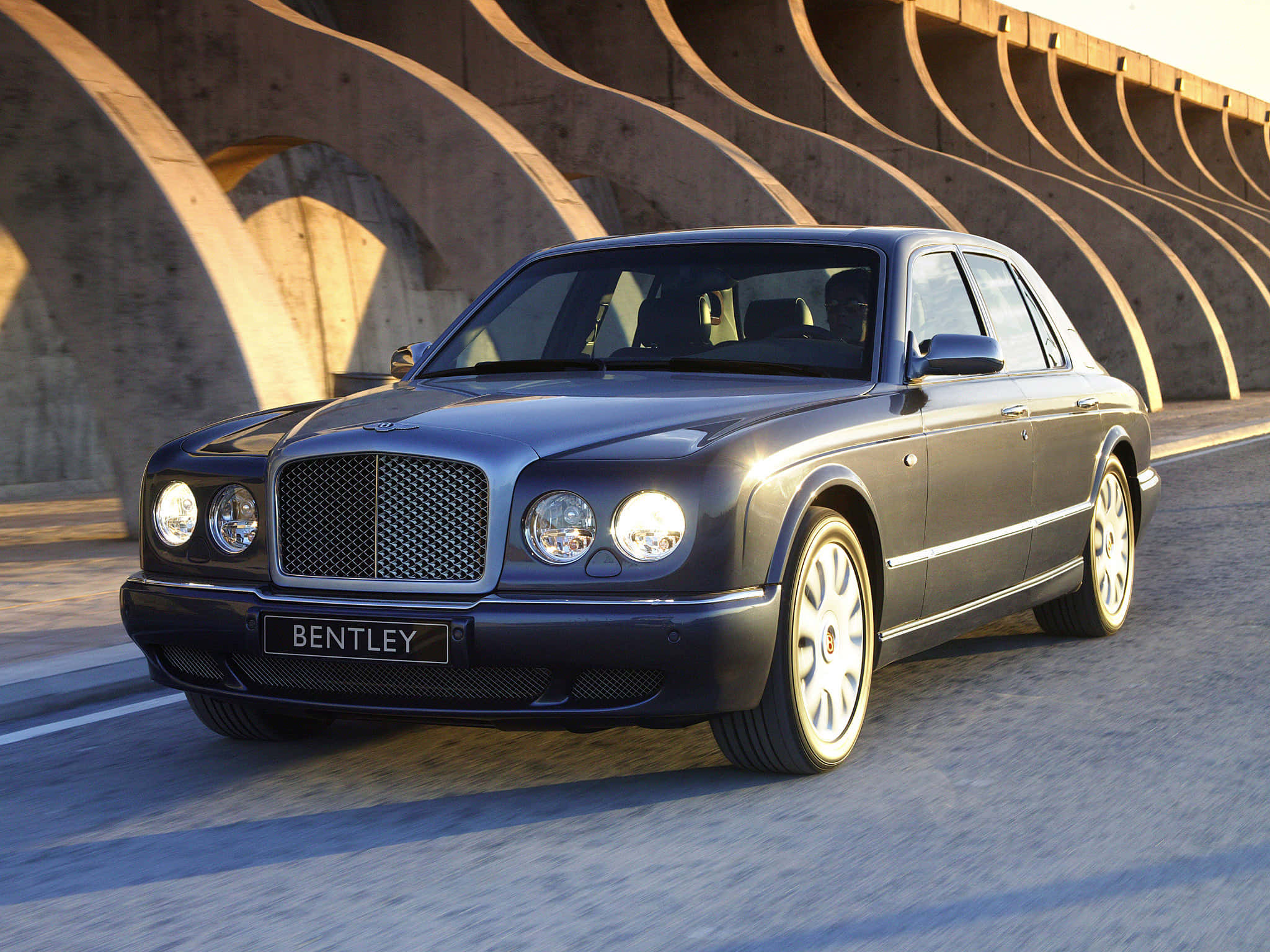 Bentley Arnage 桌布 壁紙 — 一輛時尚的深藍色賓利轎車在溫暖的黃金時段燈光下滑過彎曲的混凝土橋下。