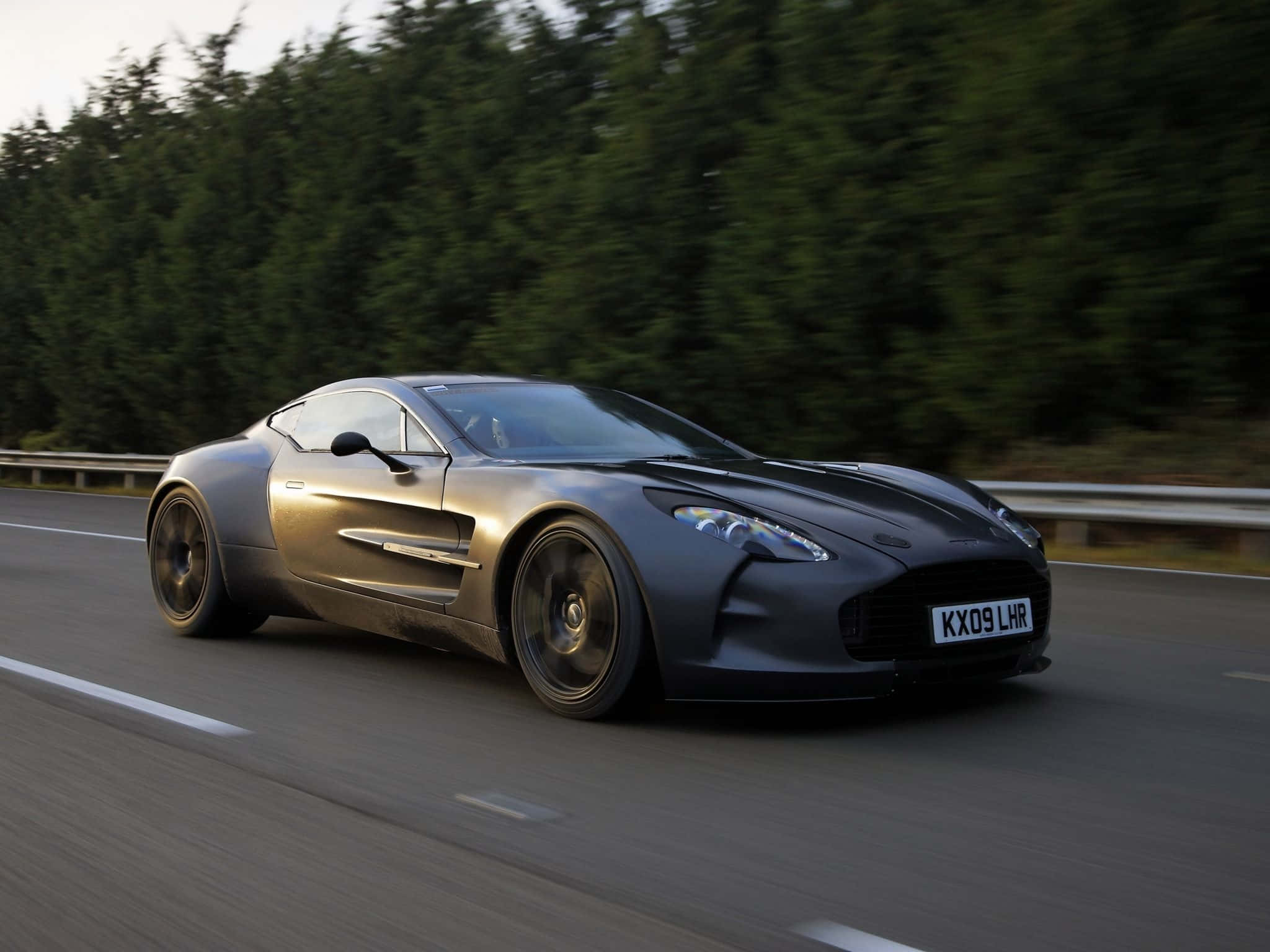 Aston Martin One-77 桌布 壁紙 — 一輛時尚的啞光灰色阿斯頓馬丁One-77在森林道路上行駛，動態模糊強調了其力量和優雅。