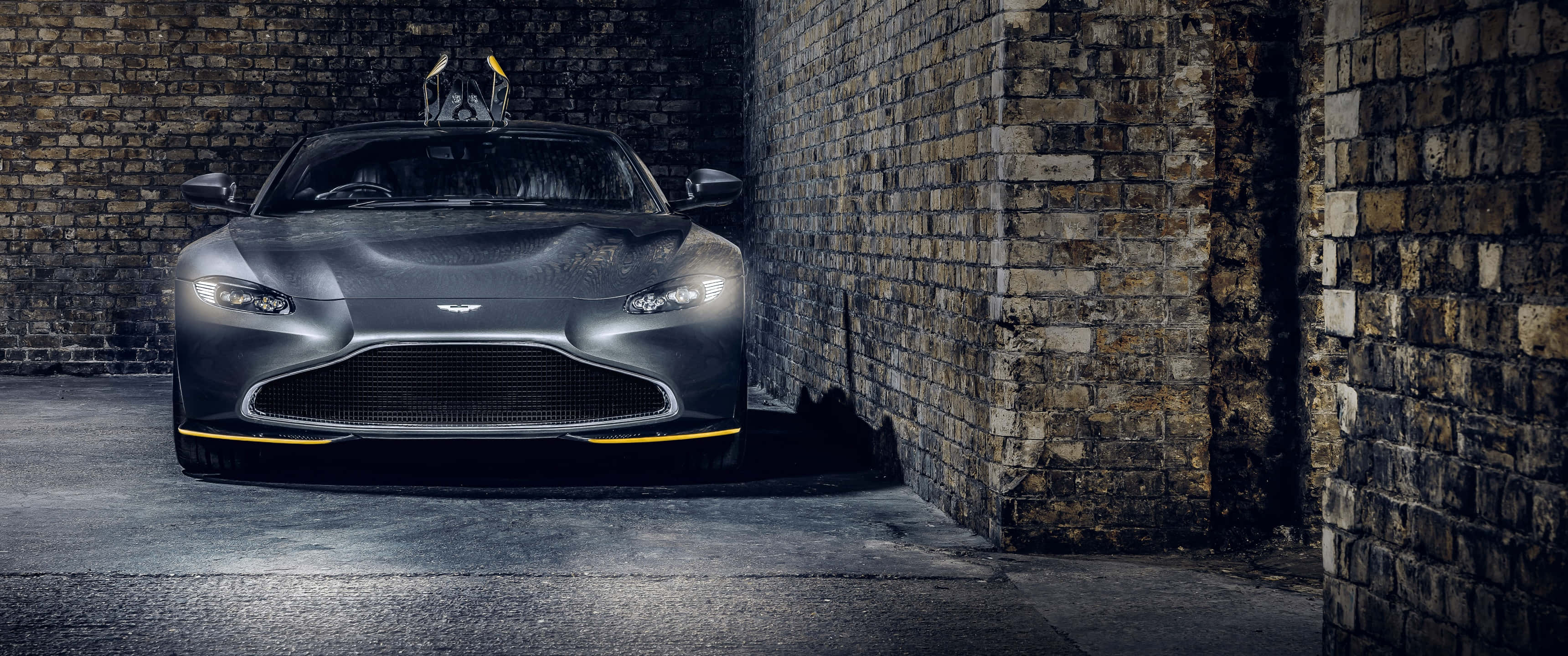 Aston Martin Vantage 桌布 壁紙 — 一輛時尚的啞光灰色阿斯頓馬丁跑車，帶有黃色點綴，停在風化的磚牆之間，光線昏暗。