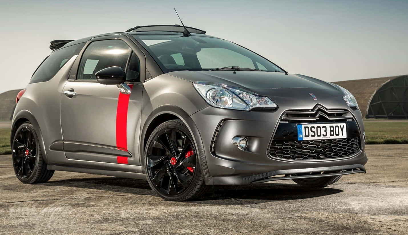  壁紙 — 一輛時尚的啞光灰色 DS 3 Racing 汽車，帶有醒目的紅色條紋和黑色輪圈，停在明亮的日光下的混凝土停車場上。