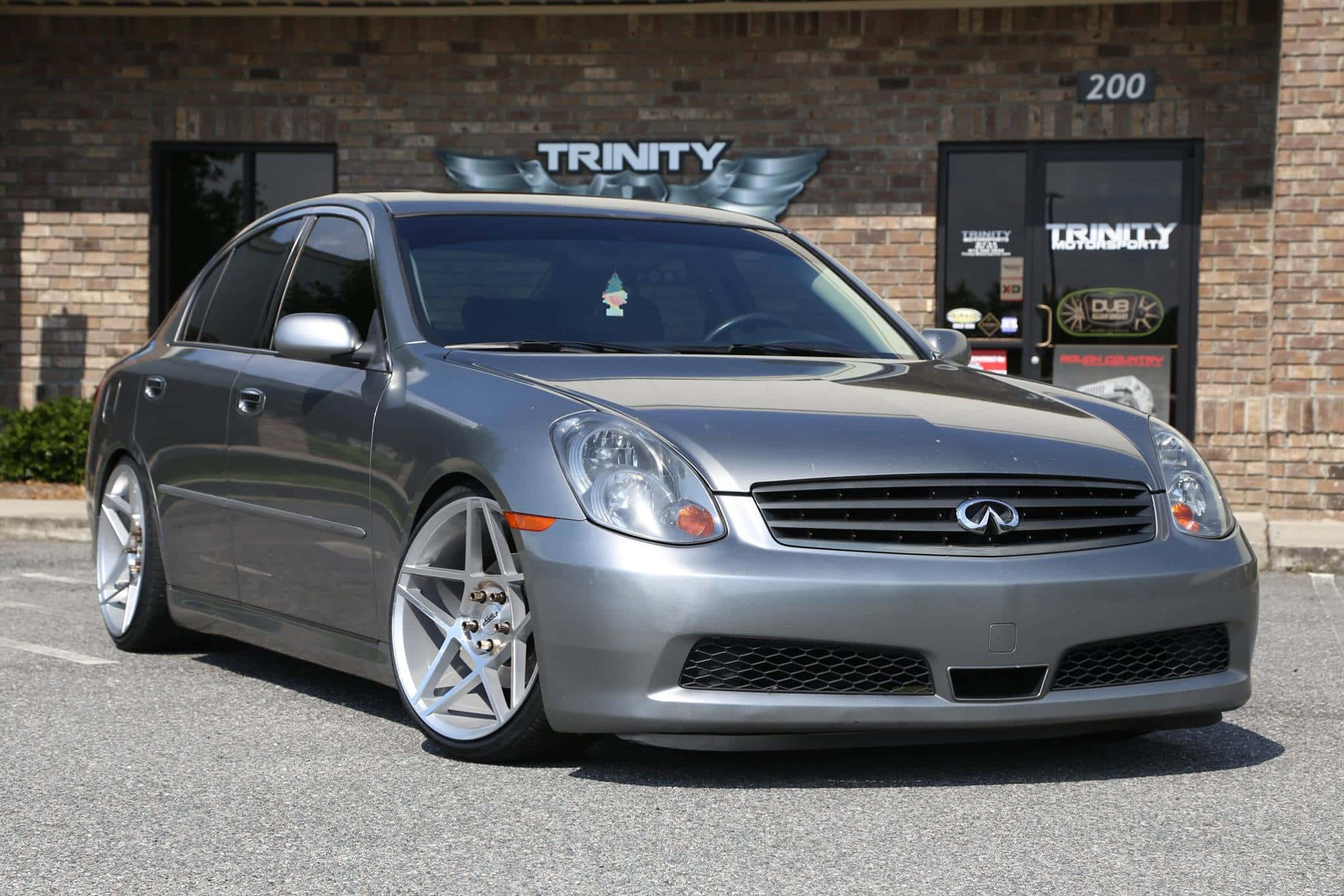 Infiniti G35 桌布 壁紙 — 一輛時尚的銀色Infiniti G35轎車，配備定制合金輪圈，停在帶有'Trinity'標誌的磚砌建築前。