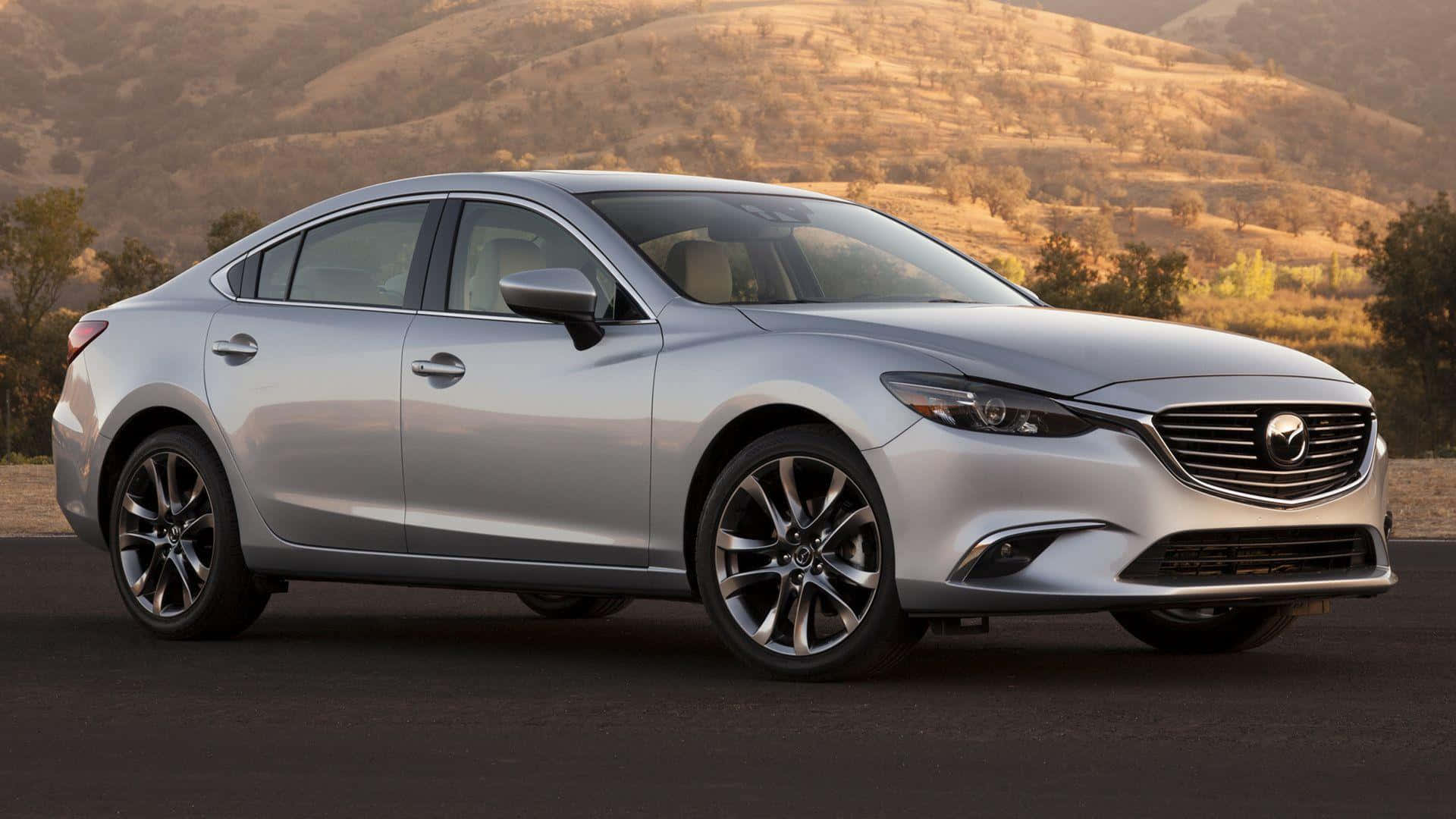 Mazda 6 桌布 壁紙 — 一輛時尚的銀色Mazda6房車停在瀝青路上，背景是溫暖陽光下的金色起伏山丘和稀疏樹木。
