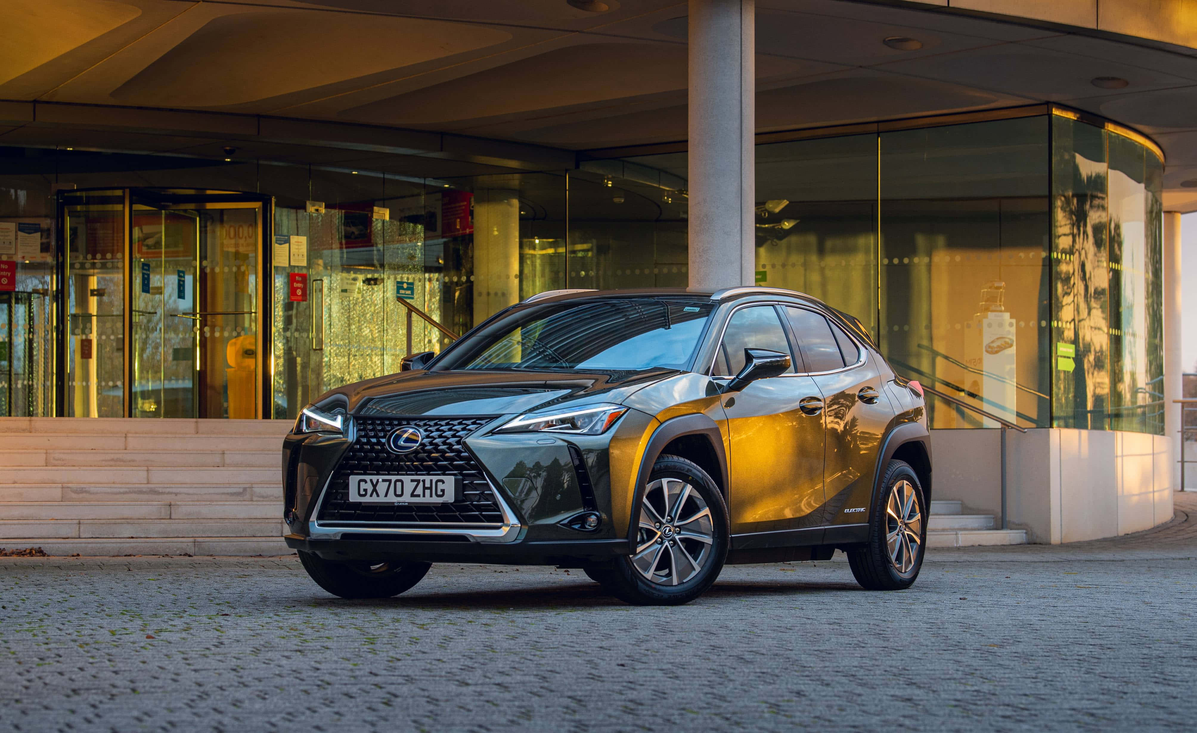 Lexus Ux 桌布 壁紙 — 一輛時尚金屬金色的Lexus UX 300e SUV停在現代玻璃建築前的鵝卵石路上，正值黃金時刻。