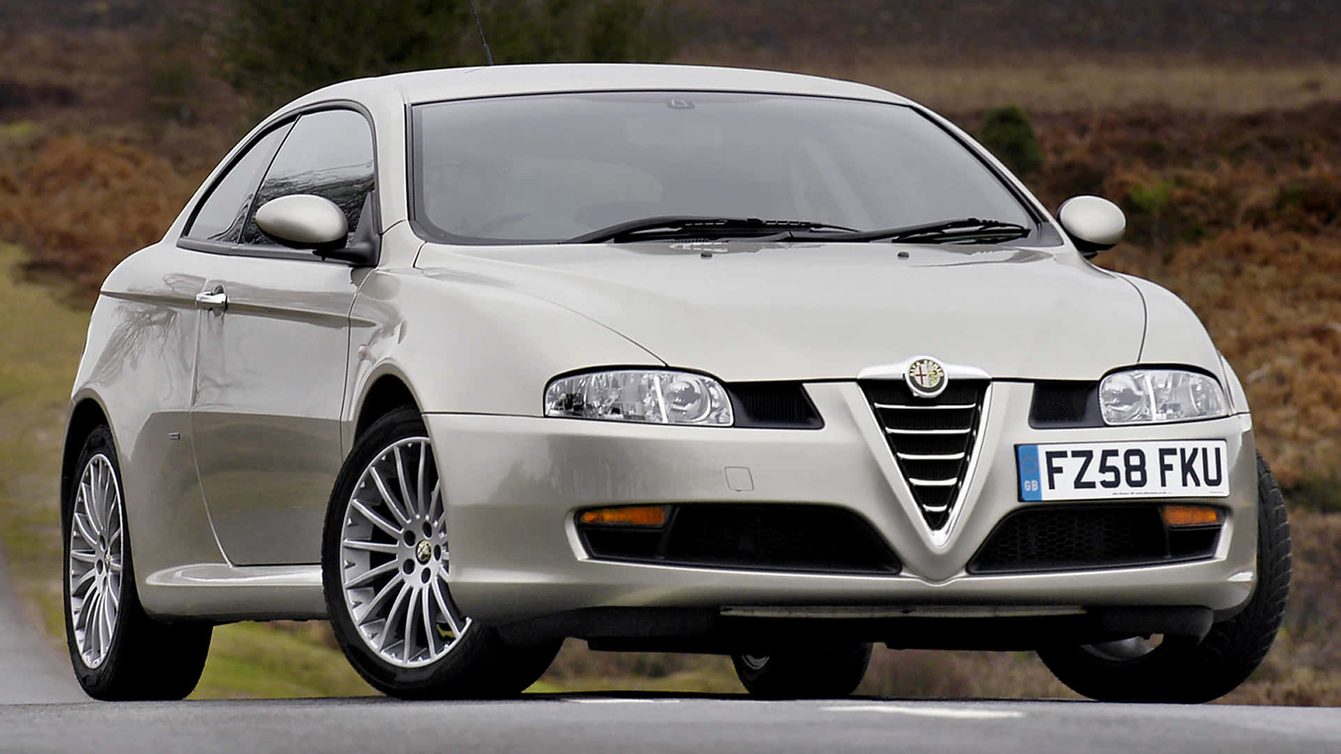 Alfa Romeo Gt 桌布 壁紙 — 一輛銀色愛快羅密歐轎跑車停在路邊，背景是長滿草的山丘。