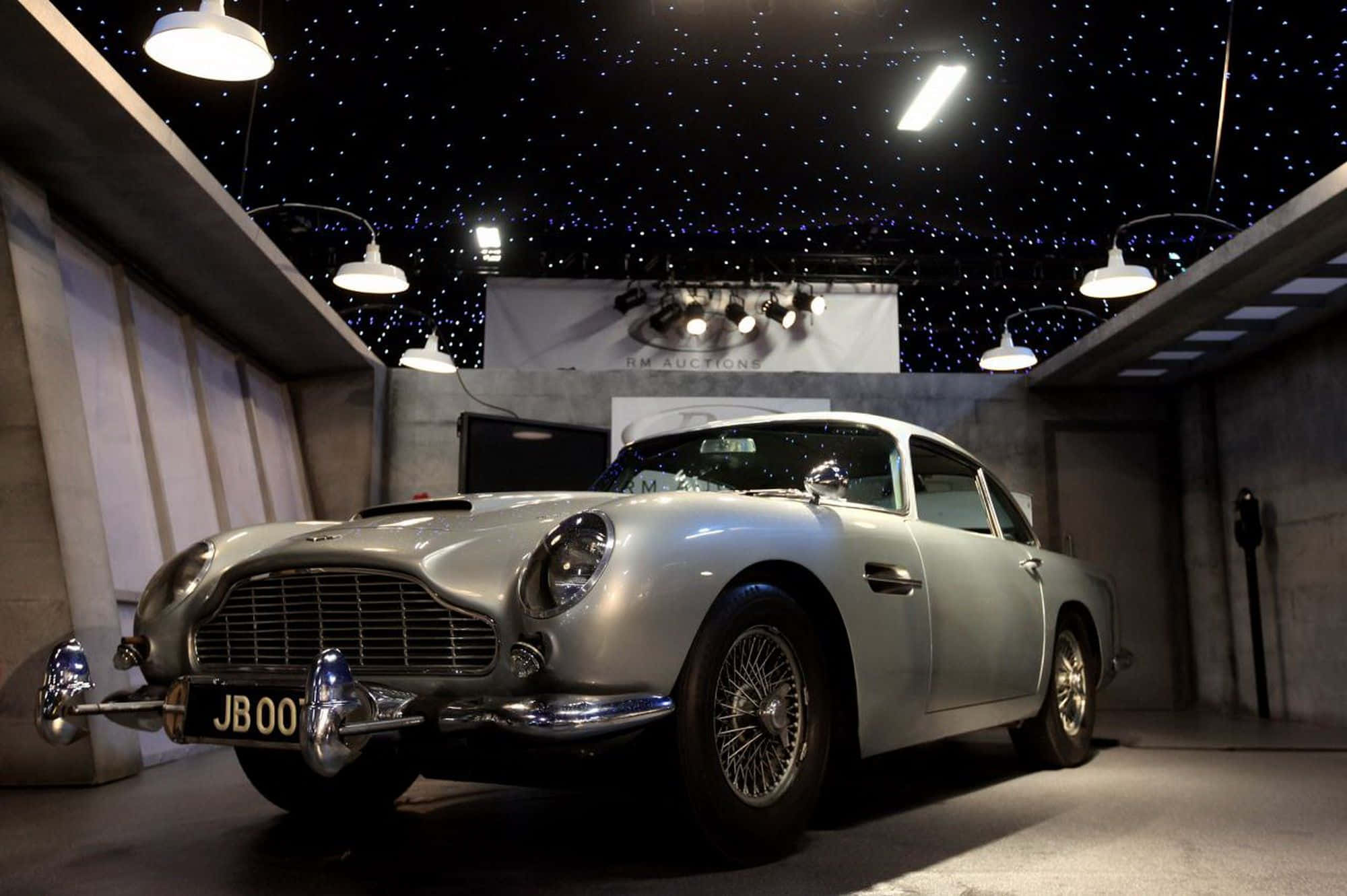 Aston Martin Db5 桌布 壁紙 — 一輛銀色Aston Martin DB5在混凝土拍賣畫廊的星空天花下，在聚光燈下閃閃發光。