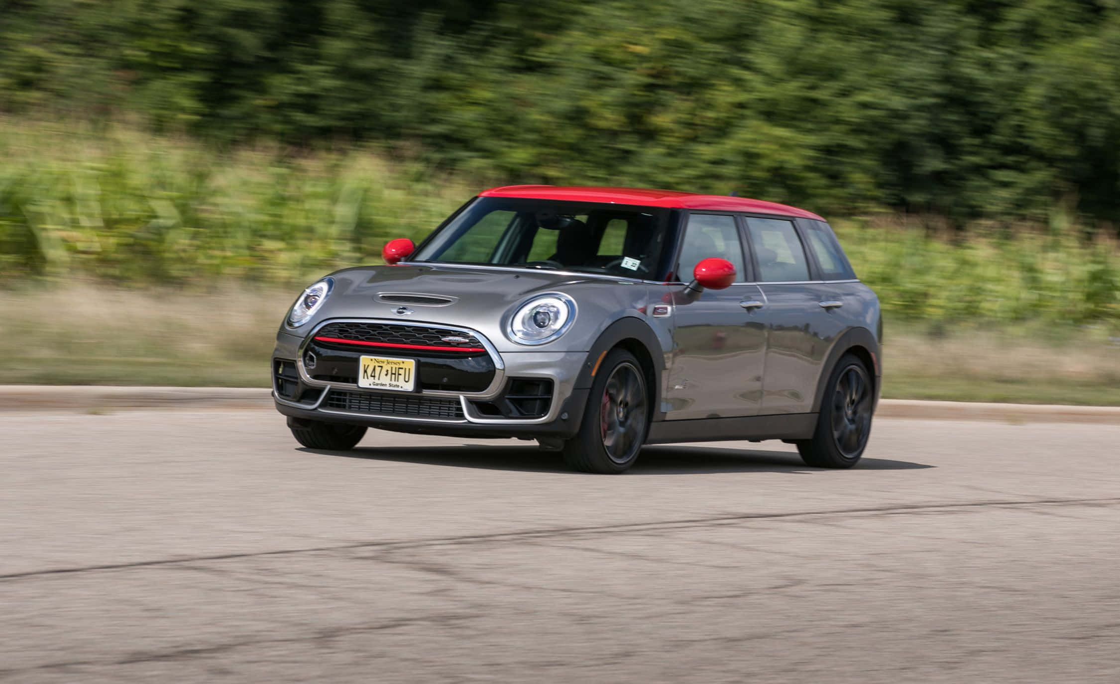 Mini Cooper S Clubman All4 桌布 壁紙 — 一輛銀色MINI Cooper，紅色車頂和後視鏡，在柏油路上飛馳，背景是模糊的綠色樹葉。