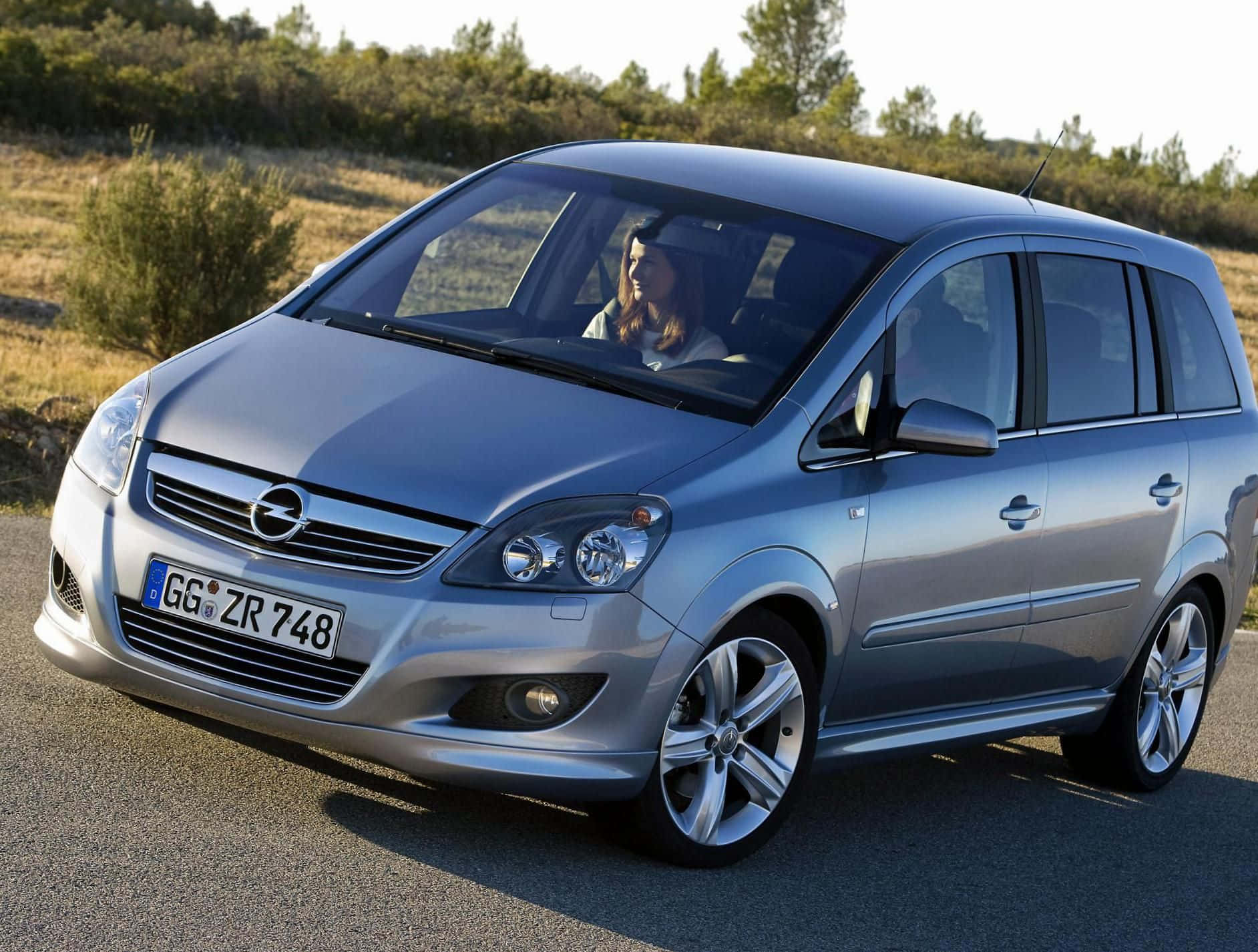 Opel Zafira 桌布 壁紙 — 一輛銀色歐寶Zafira停在瀝青路上，透過擋風玻璃可以看到一名女司機，背景是乾燥的灌木叢景觀。