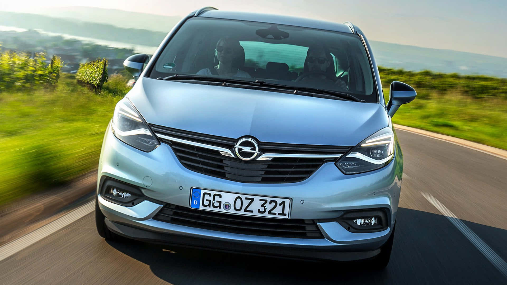 Opel Zafira 桌布 壁紙 — 一輛銀色歐寶Zafira在陽光明媚的道路上快速行駛，模糊的綠色山丘飛馳而過。