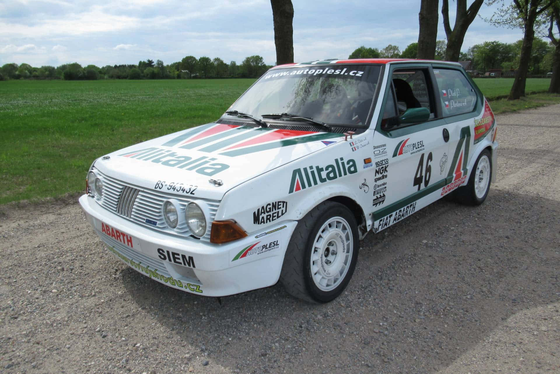 Fiat Ritmo 桌布 壁紙 — 一輛白色Fiat Abarth拉力賽車，帶有Alitalia塗裝，停在碎石路上，旁邊是草地，天空多雲。