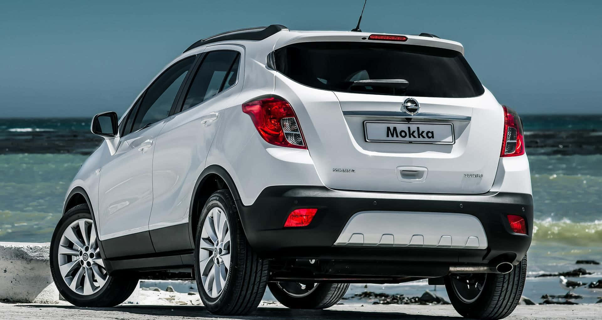 Opel Mokka 桌布 壁紙 — 一輛白色Opel Mokka SUV停泊在岩石海灘上，海浪在清澈藍天下拍打岸邊。