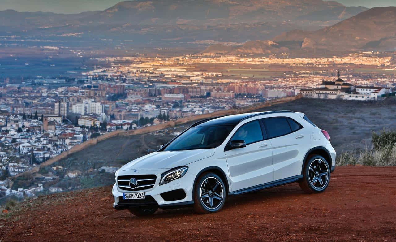 Mercedes Benz Gla-class 桌布 壁紙 — 一輛白色平治GLA-Class SUV停泊在紅色泥土觀景點上，俯瞰著沐浴在金色夕陽下的廣闊城市和山脈。