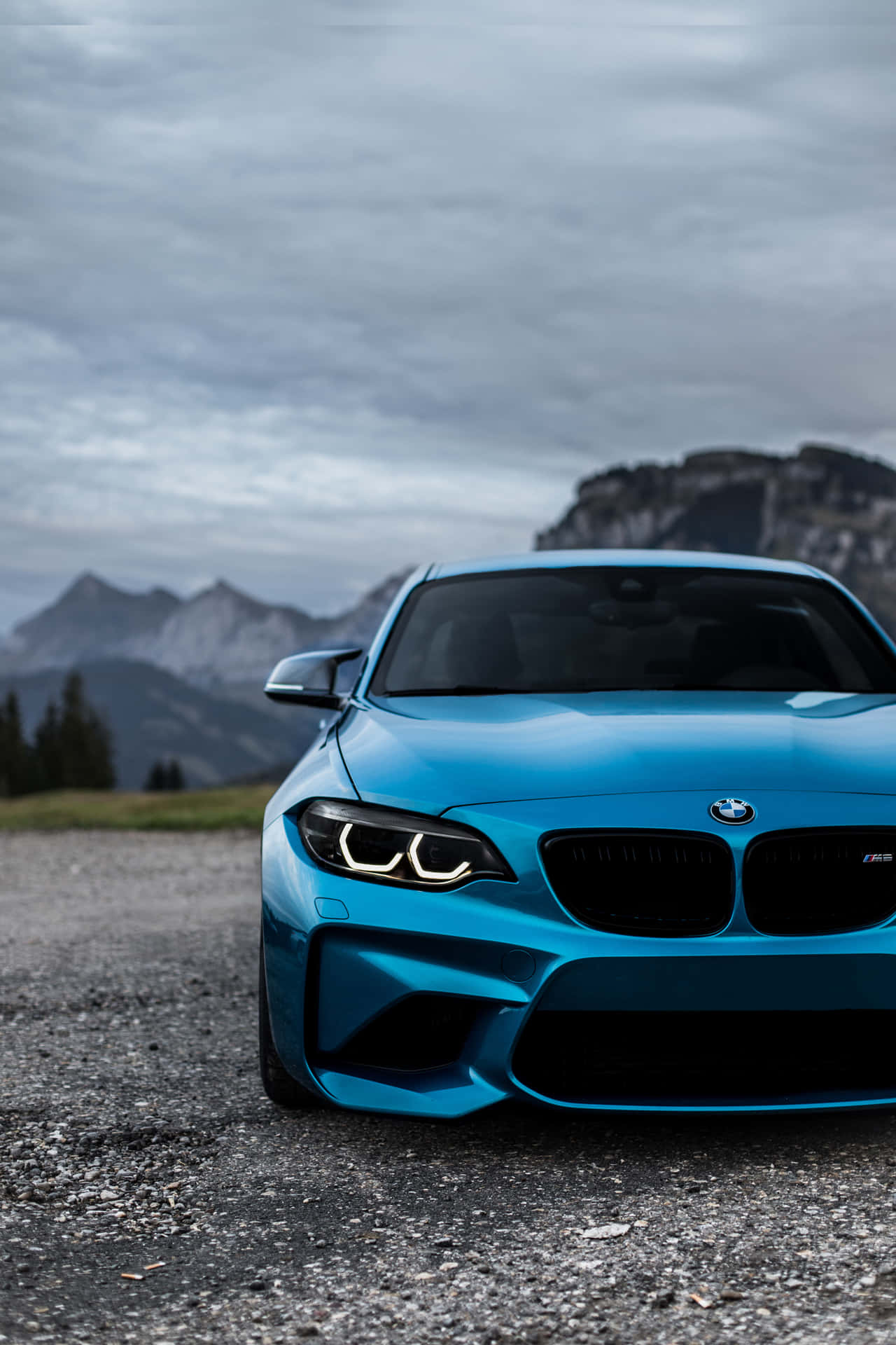 藍色BMW M2跑車停在礫石上，靠近山脈，天空多雲。