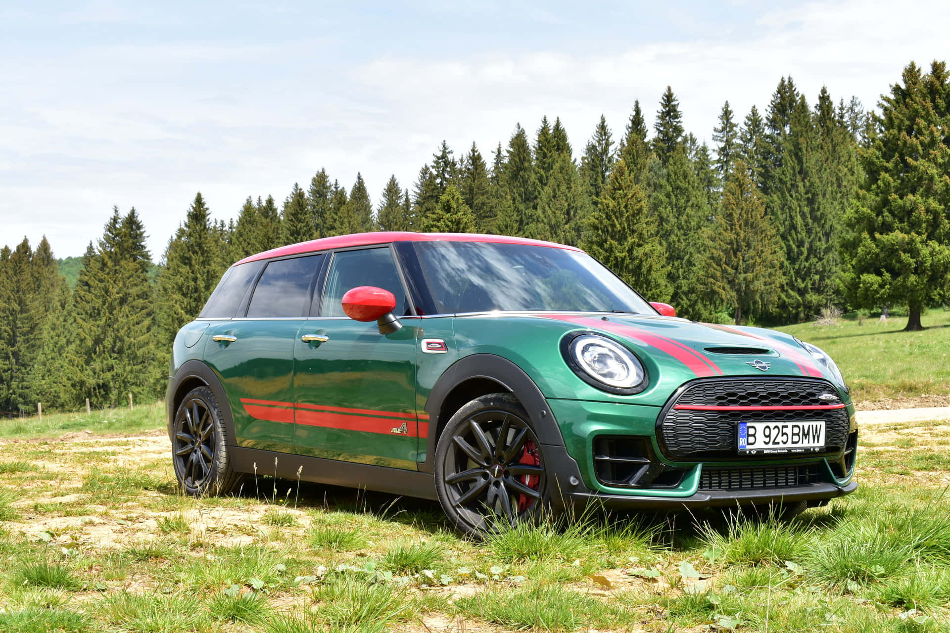 Mini Cooper S Clubman All4 桌布 壁紙 — 一輛充滿活力的綠色MINI Cooper，帶有紅色點綴，停在草地上，周圍是茂密的松樹，天空明亮。