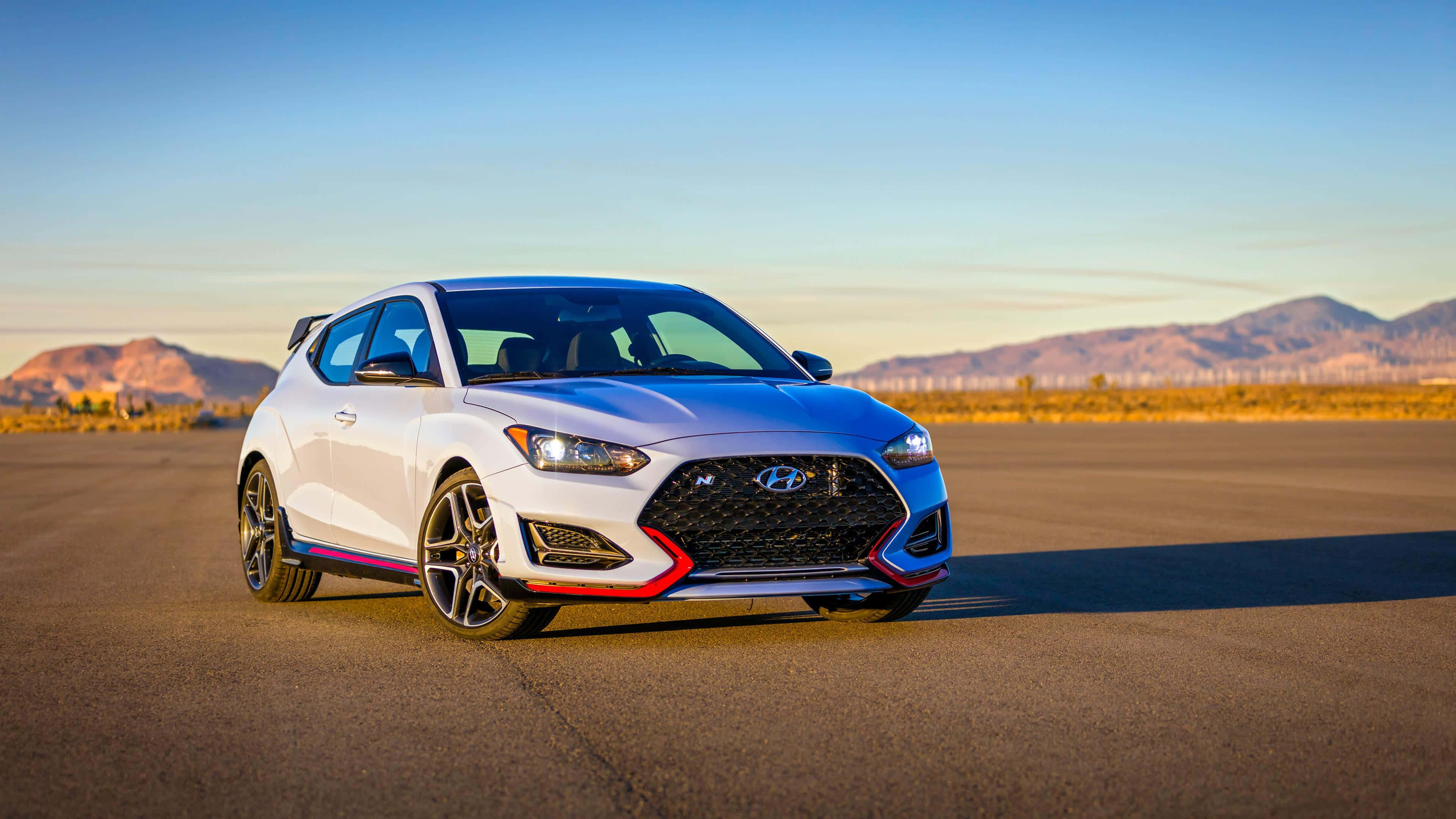 Hyundai Veloster 桌布 壁紙 — 一輛帶有紅色飾邊的白色現代Veloster N跑車，停在空蕩的瀝青路上，背景是日落時分的沙漠山脈。