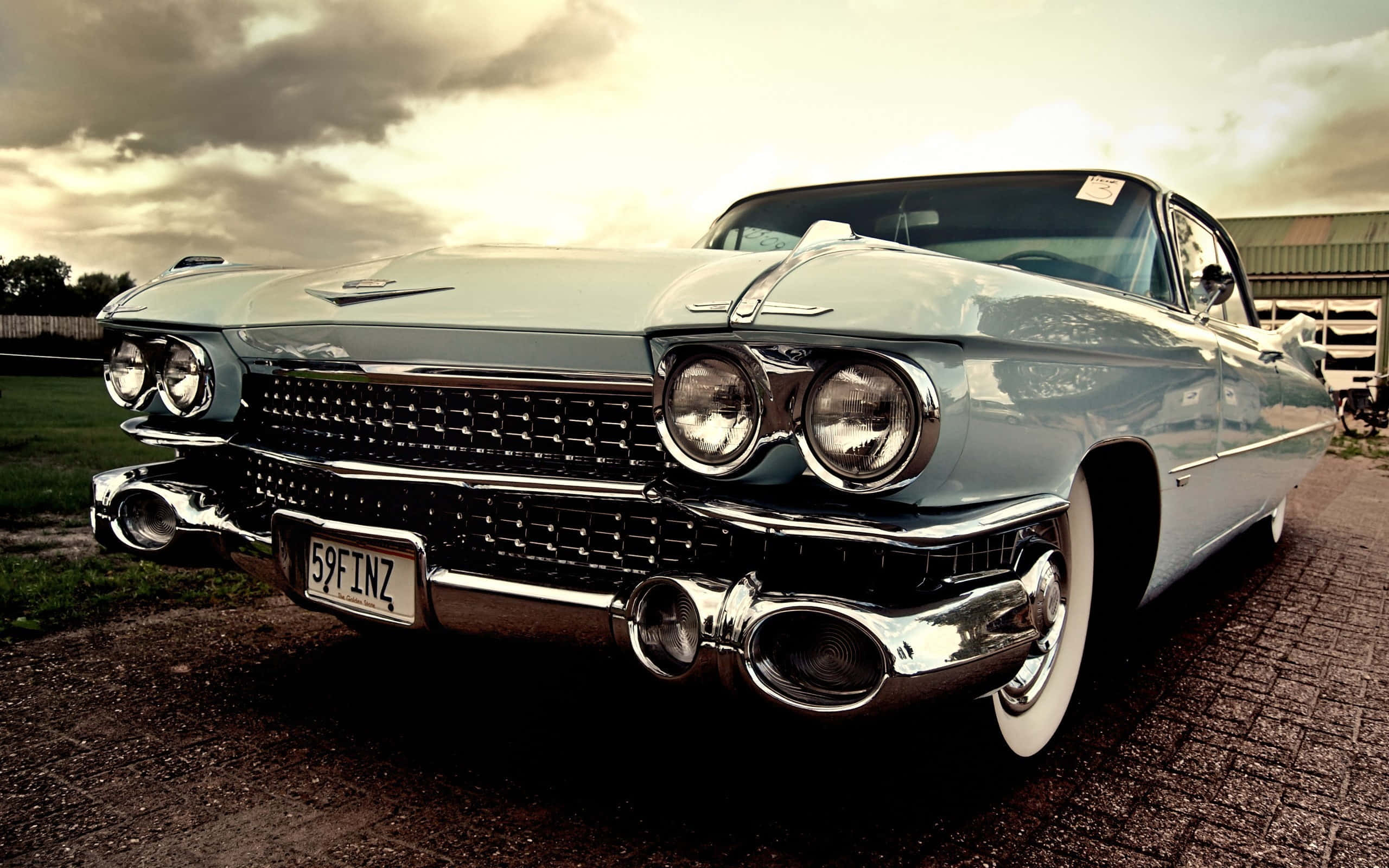 Cadillac Deville 桌布 壁紙 — 一輛帶有鍍鉻細節的復古淺藍色凱迪拉克，在戲劇性的多雲天空下，在鋪砌的車道上閃閃發光。