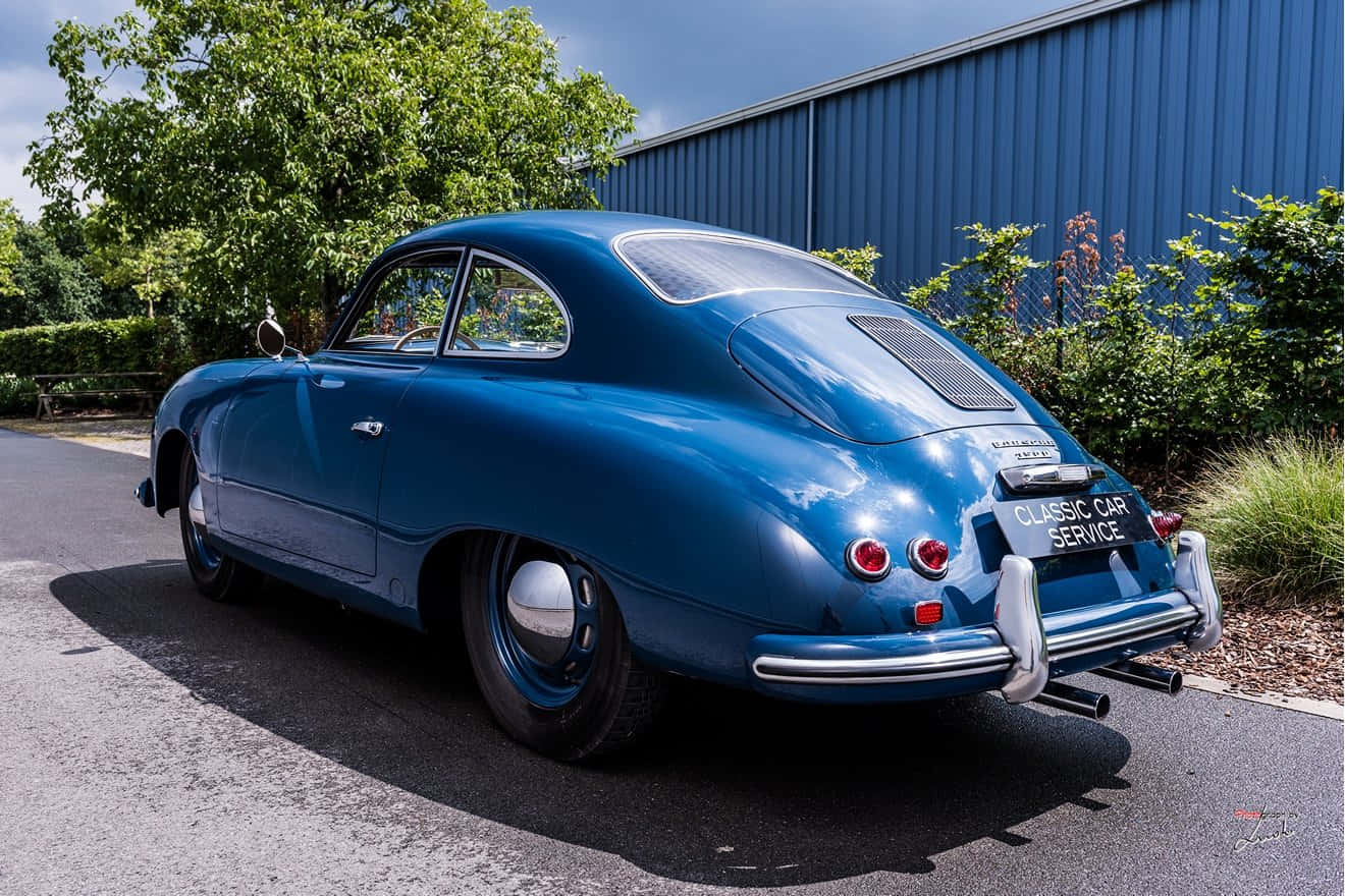 Porsche 356 桌布 壁紙 — 一輛光亮的藍色經典保時捷356停在瀝青路上，旁邊有綠植和藍色建築，在明亮的日光下。