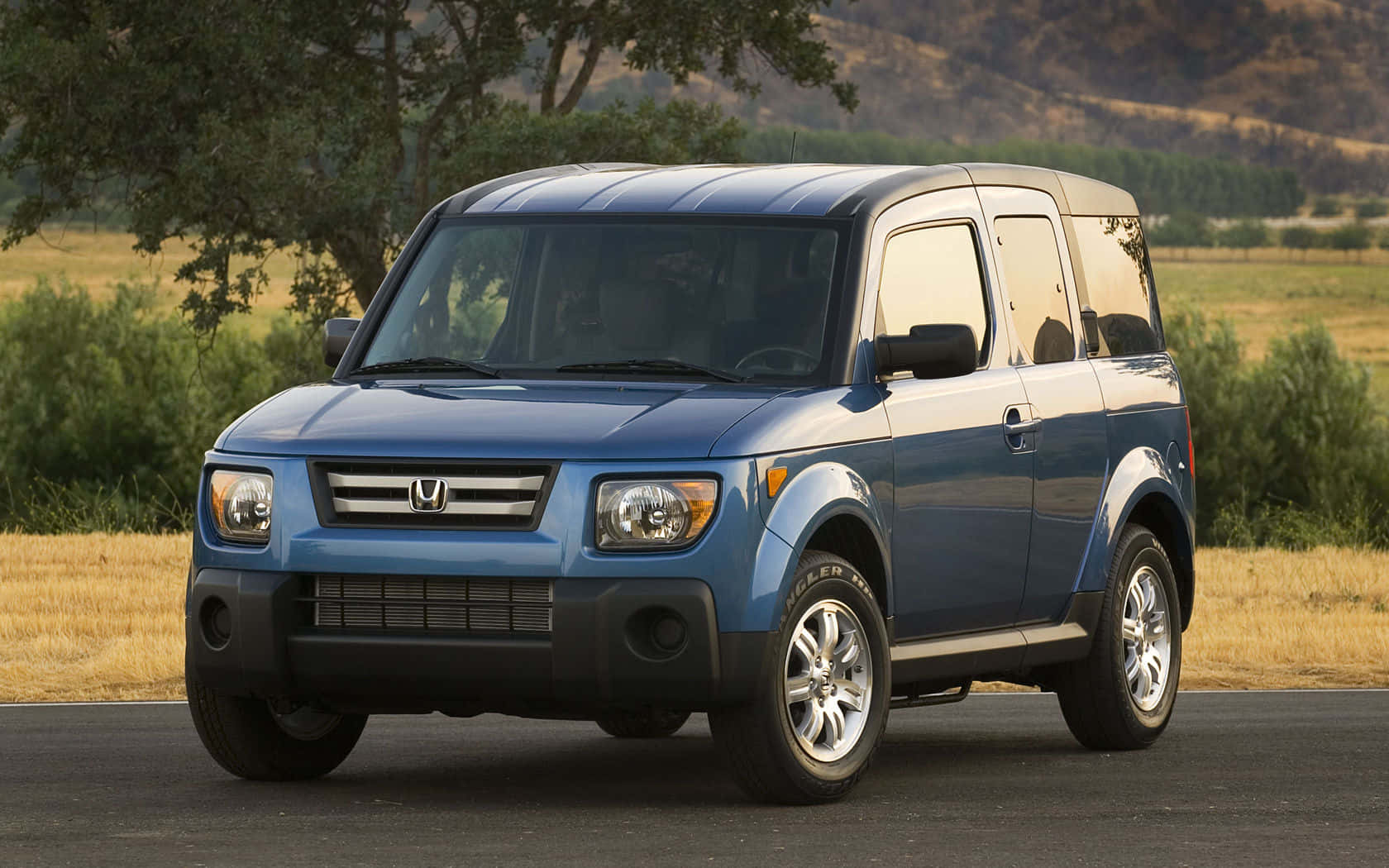 Honda Element 桌布 壁紙 — 一輛藍色本田 Element SUV 停在瀝青路上，背景有樹木和山丘，沐浴在溫暖的陽光下。