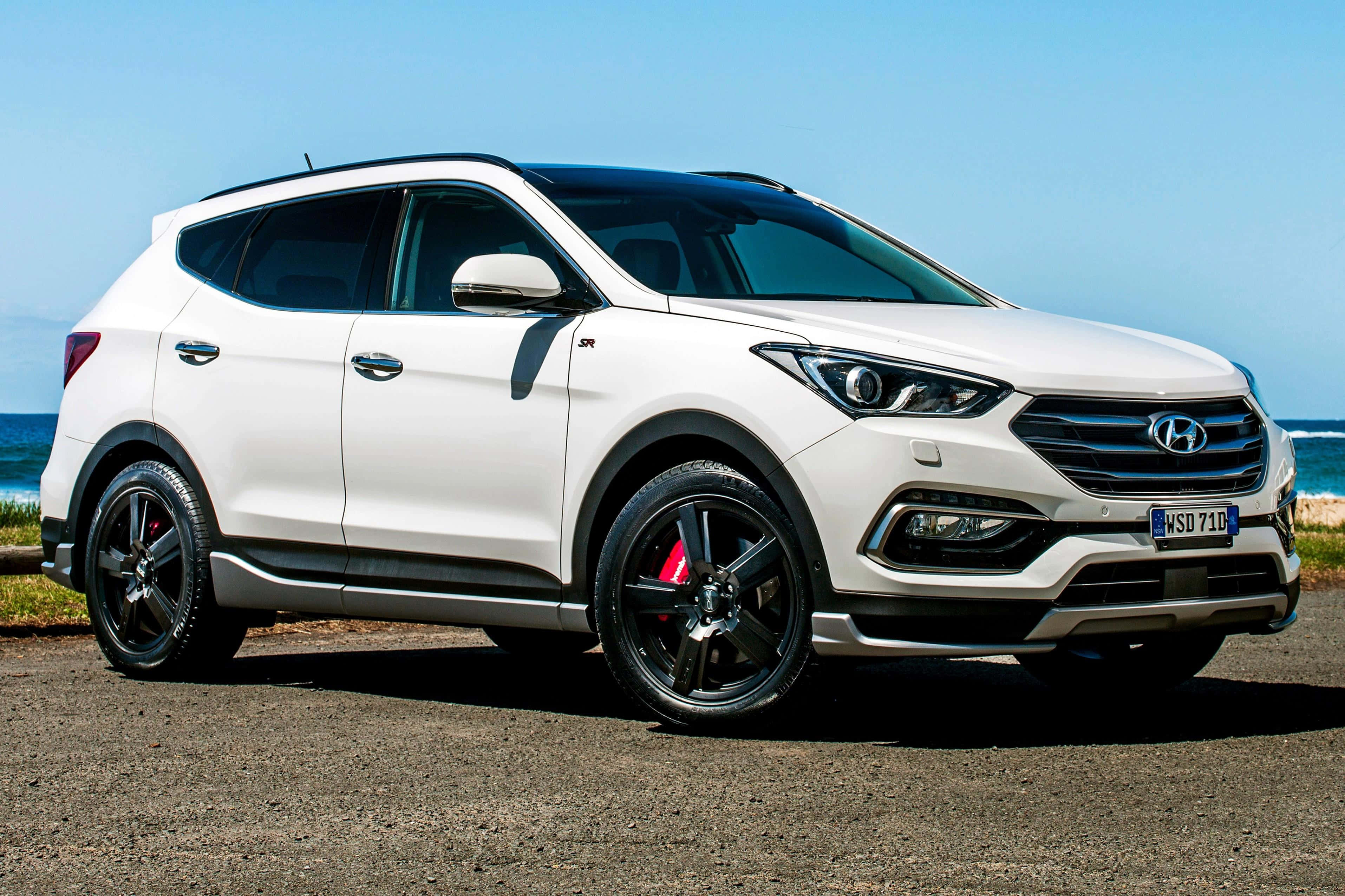 Hyundai Tucson 桌布 壁紙 — 一輛時尚的白色現代Santa Fe SUV，配備黑色輪圈和紅色制動卡鉗，停在藍天白雲下的海邊礫石上。