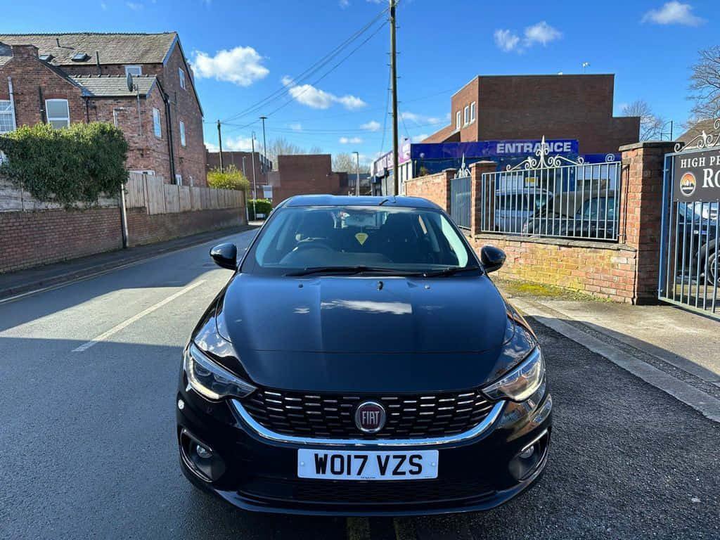 Fiat Tipo 桌布 壁紙 — 一輛時尚的黑色Fiat Punto停泊在陽光普照的街道上，旁邊是磚砌建築和一個有閘門的入口。