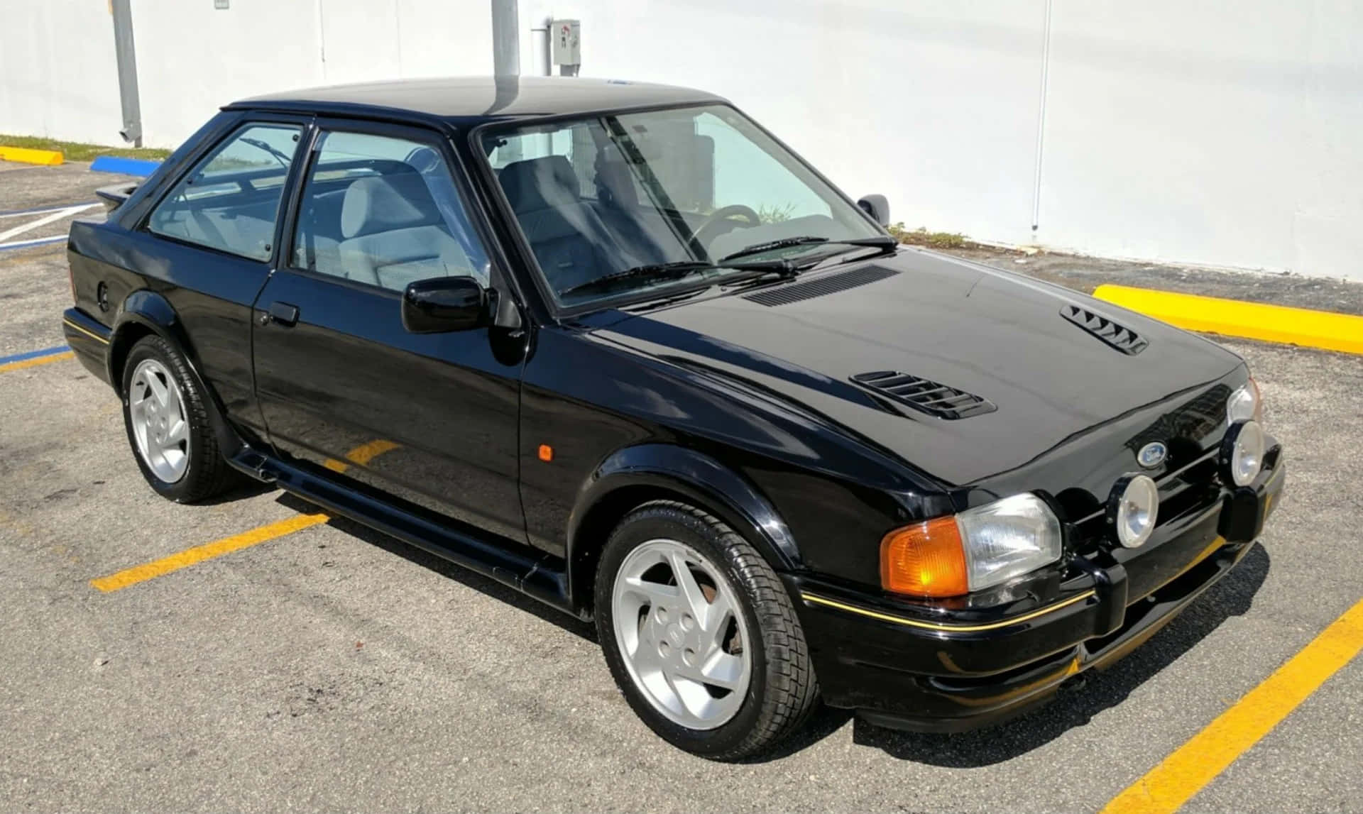 Ford Escort 桌布 壁紙 — 一輛時尚的黑色福特Sierra Cosworth跑車停泊在陽光普照的瀝青停車場，地上有黃線。